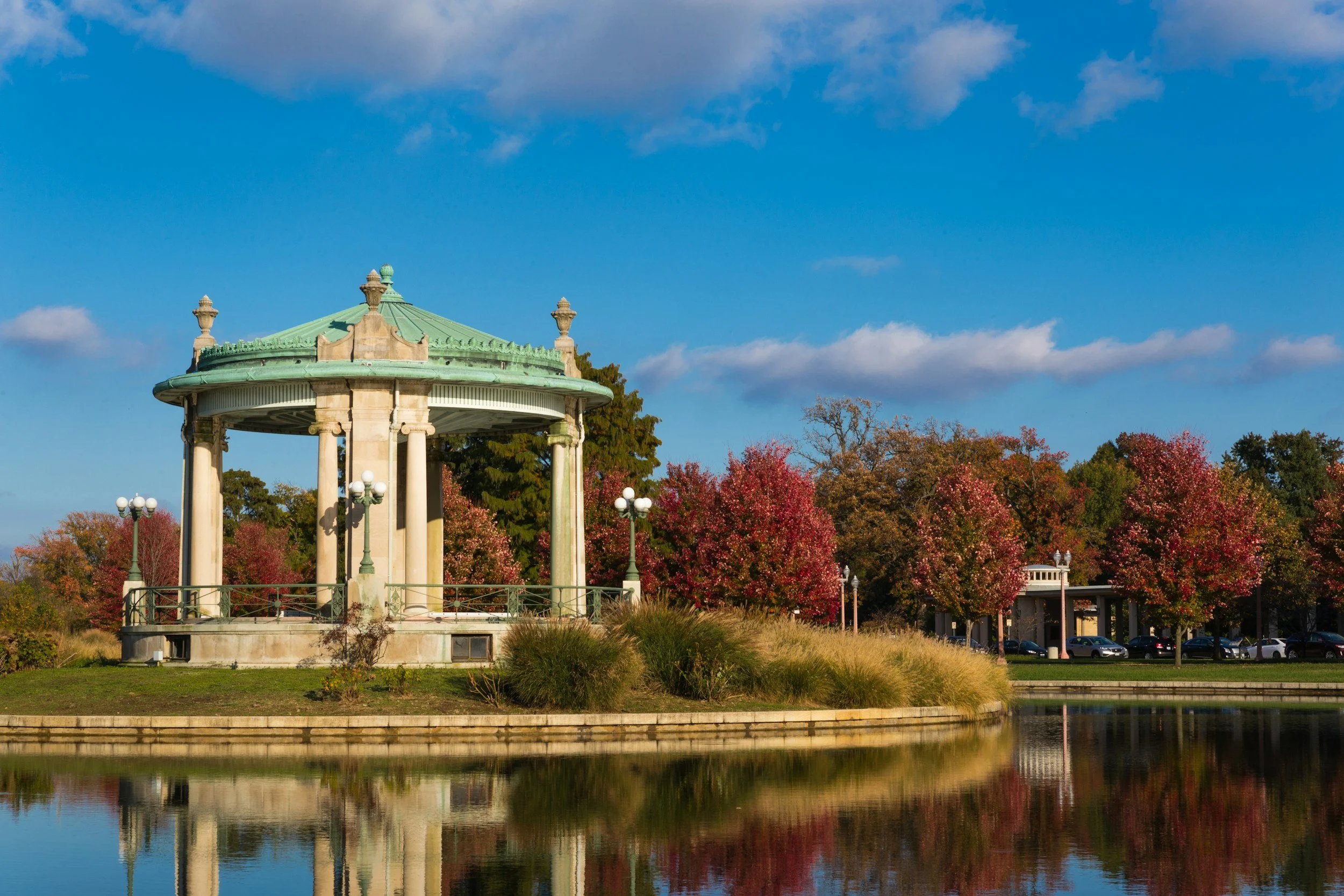 A classical gazebo with green roof and ornamental decor, located near a body of water with reflection, surrounded by autumn-colored trees and a partly cloudy sky.