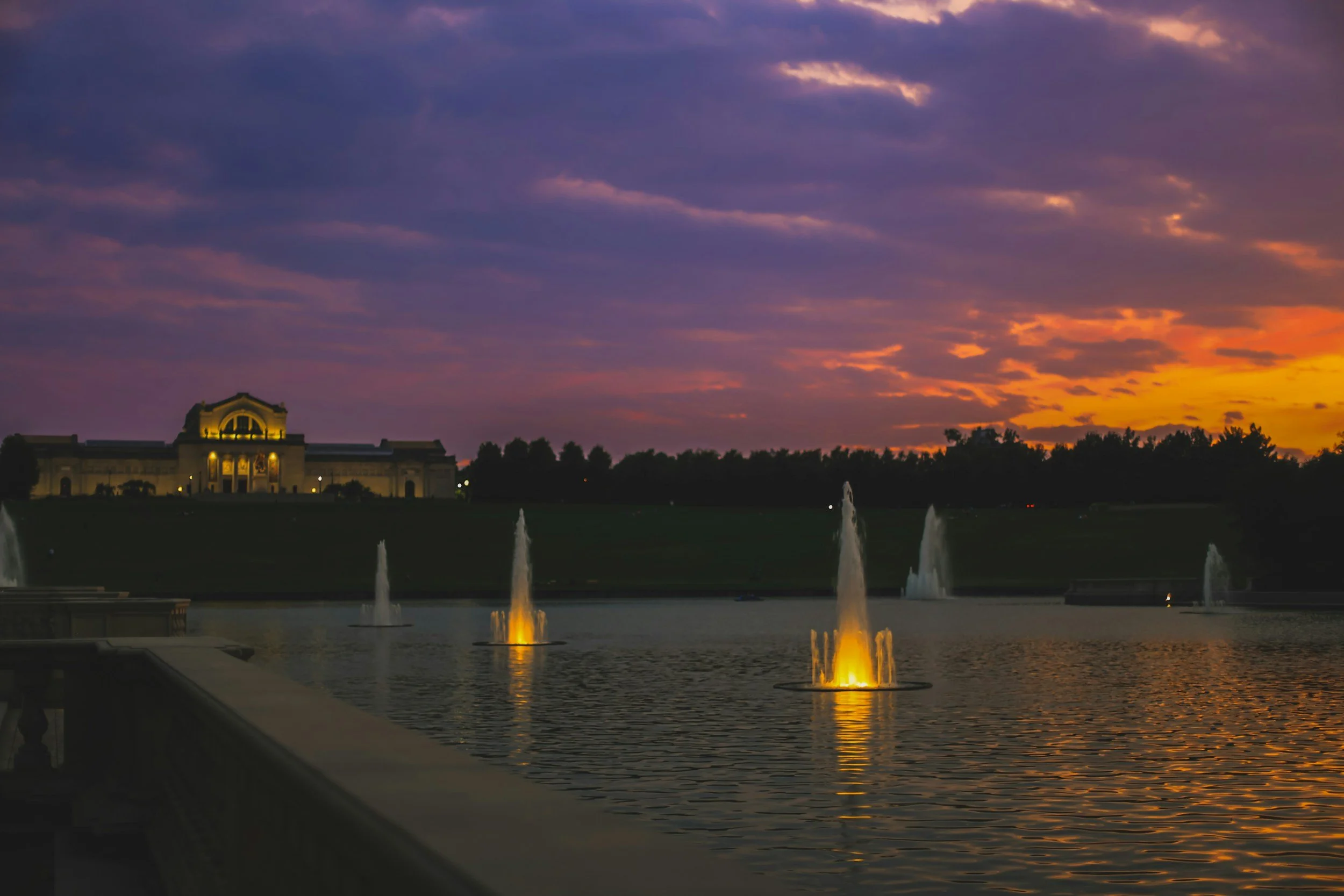 A large building with classical architecture under a colorful sunset sky, with fountains in a reflecting pool in the foreground.
