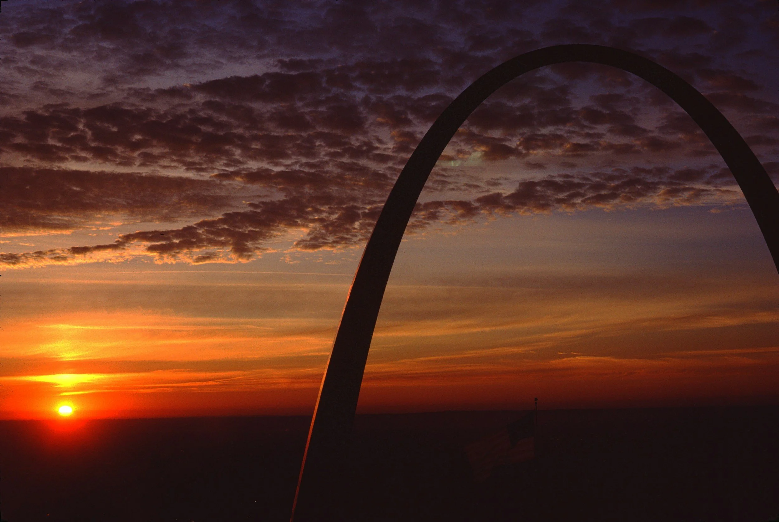 Sunset over the horizon with a partly cloudy sky and the Gateway Arch in St. Louis silhouetted against the colorful sky.