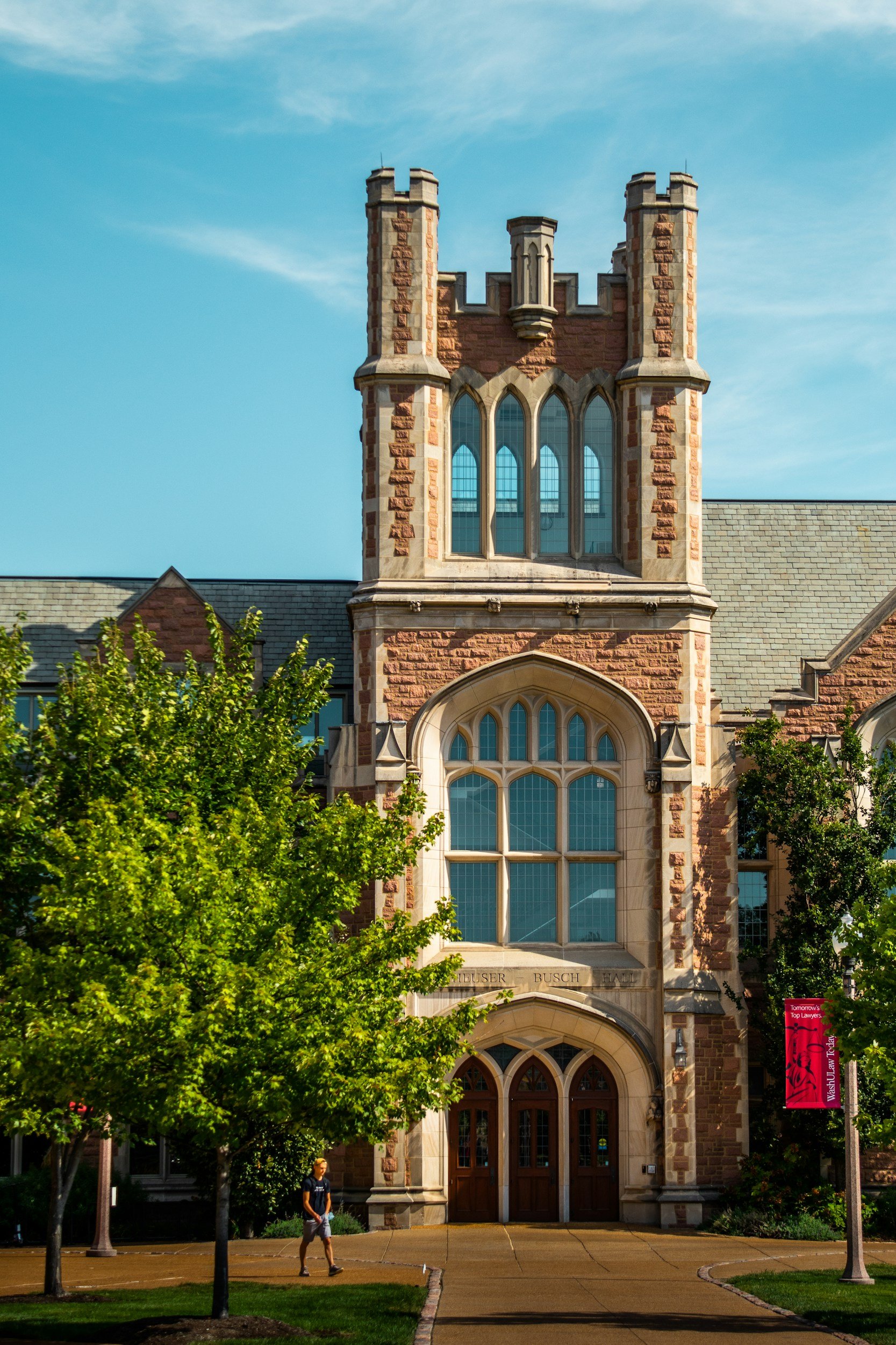 A historic red brick building with gothic windows and a clock tower, surrounded by green trees and a sidewalk with a person walking.