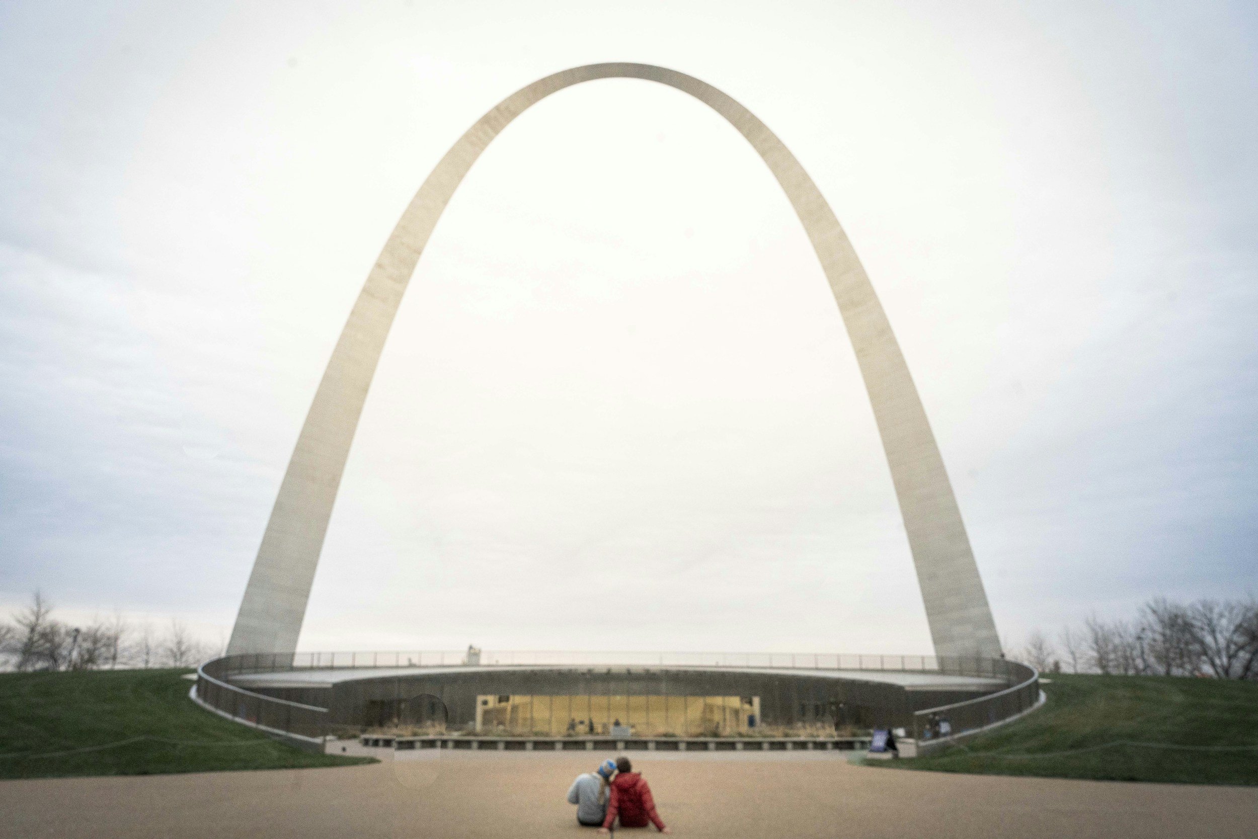 The Gateway Arch, a large stainless steel arch structure in St. Louis, Missouri, viewed against a cloudy sky. Two children are sitting on the ground in the foreground.