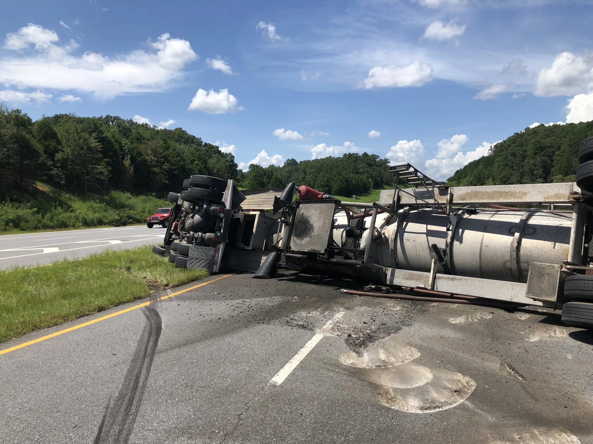 A large truck accident on a highway with a tanker truck overturned on the side of the road, spilling a liquid onto the pavement. A red vehicle and a white vehicle are visible in the background, a heavy duty tow truck 50T rotator is on the way