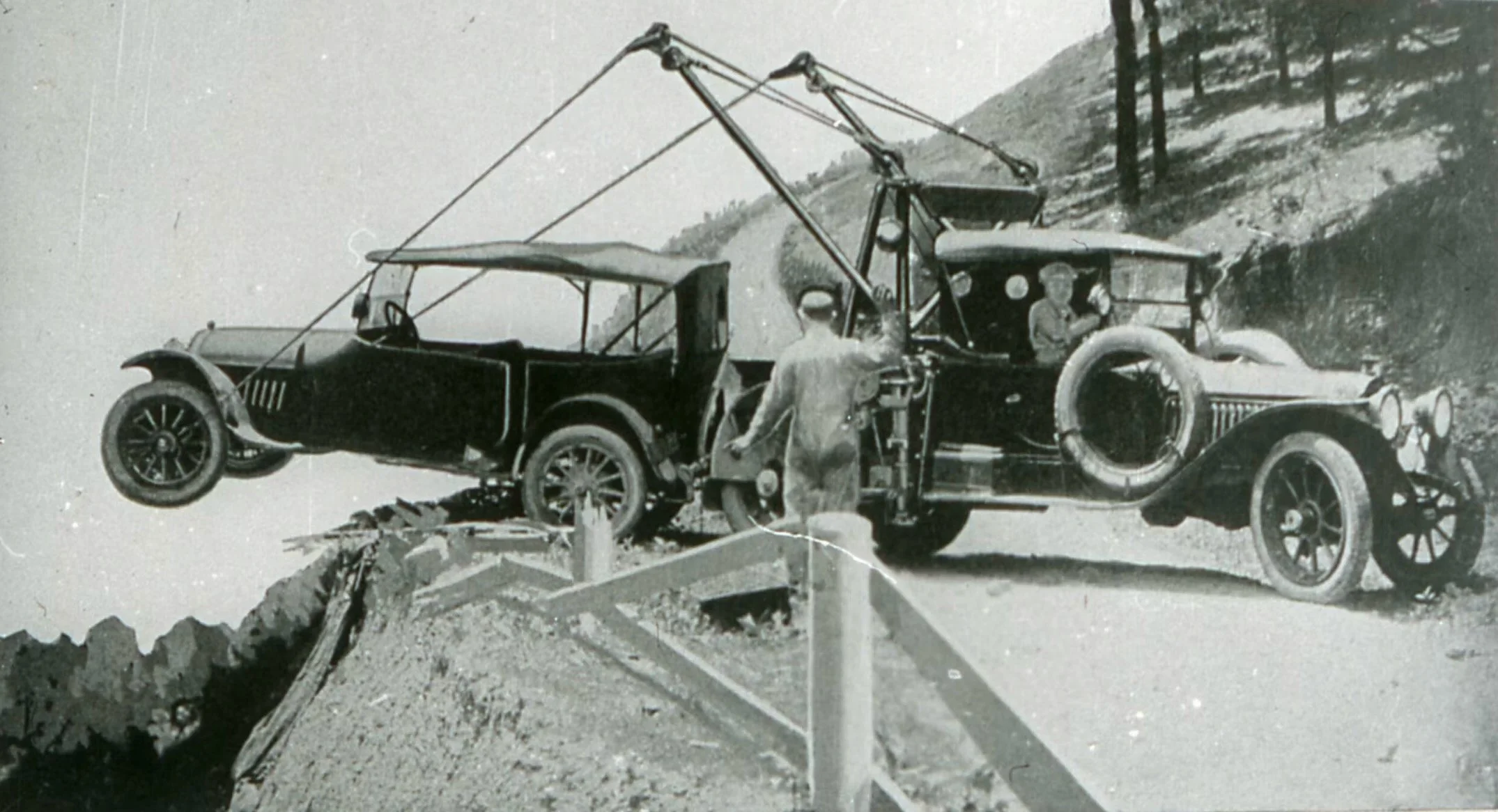 A black-and-white photograph of an early vintage car being lifted or moved, with two men standing nearby, one man is standing on the ground and the other is inside the vehicle. The setting appears to be around lookout mountain