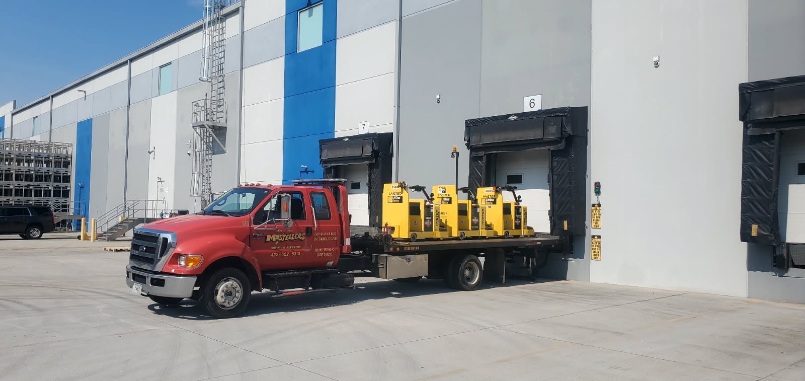 A red tow truck conducting a load transfer parked outside a warehouse with yellow and black pallet jacks on its flatbed. The warehouse has loading bays numbered 5, 6, and 7.