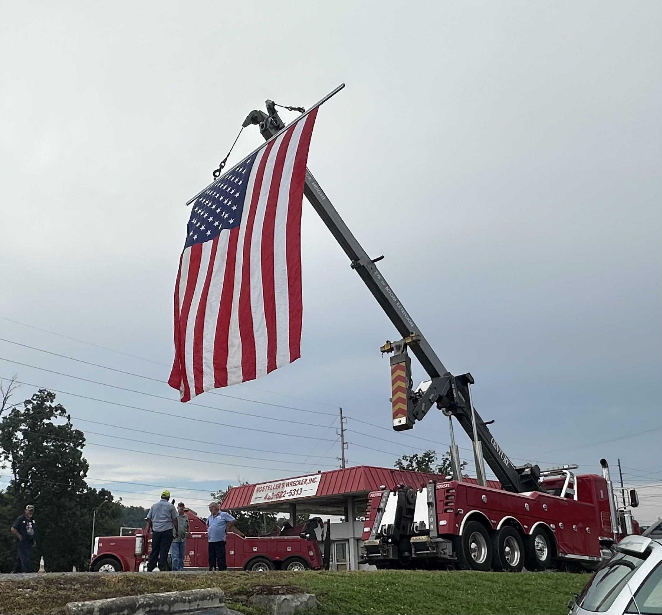 A fire truck with a mechanical lift raising an American flag high in the sky against a cloudy sky. Several people are standing near the truck, and power lines are visible in the background.