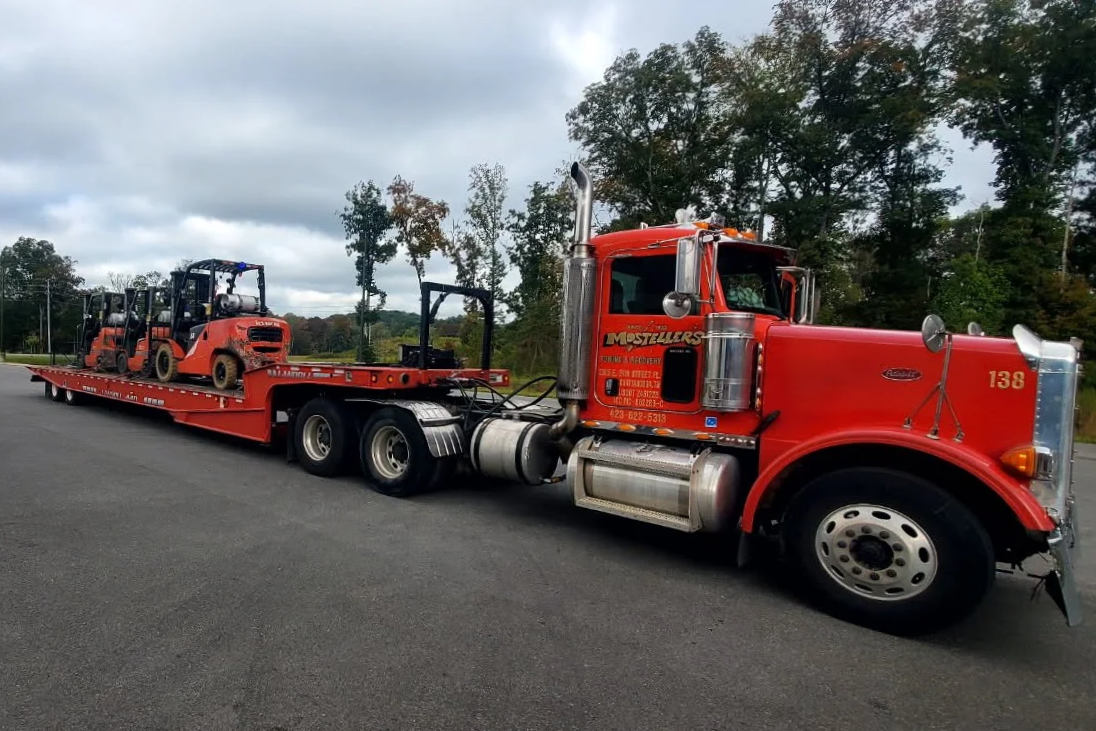 Red flatbed tow truck carrying two small orange heavy equipment fork lift vehicles on an overcast day.