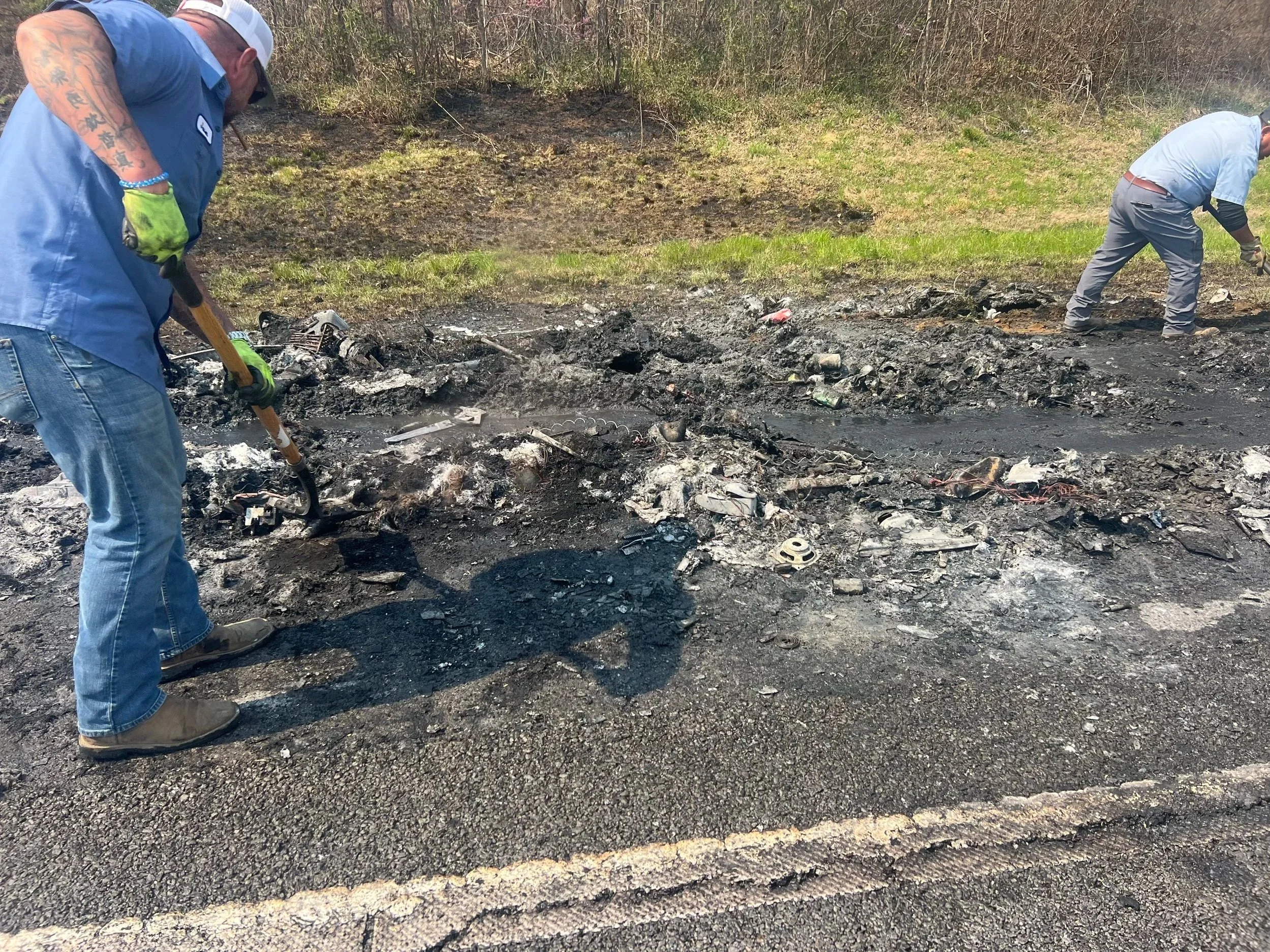 Two tow truck operators are digging through the remains of a burned area, clearing ash and debris from what appears to be a burned forest or grassland, along side the highway