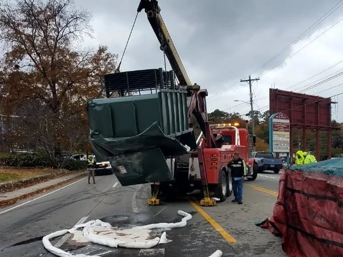 A large crane truck is lifting a green cart from the middle of a road after a crash. Debris, including a crumpled white object and a red vehicle, is scattered on the road. Emergency personnel are present.