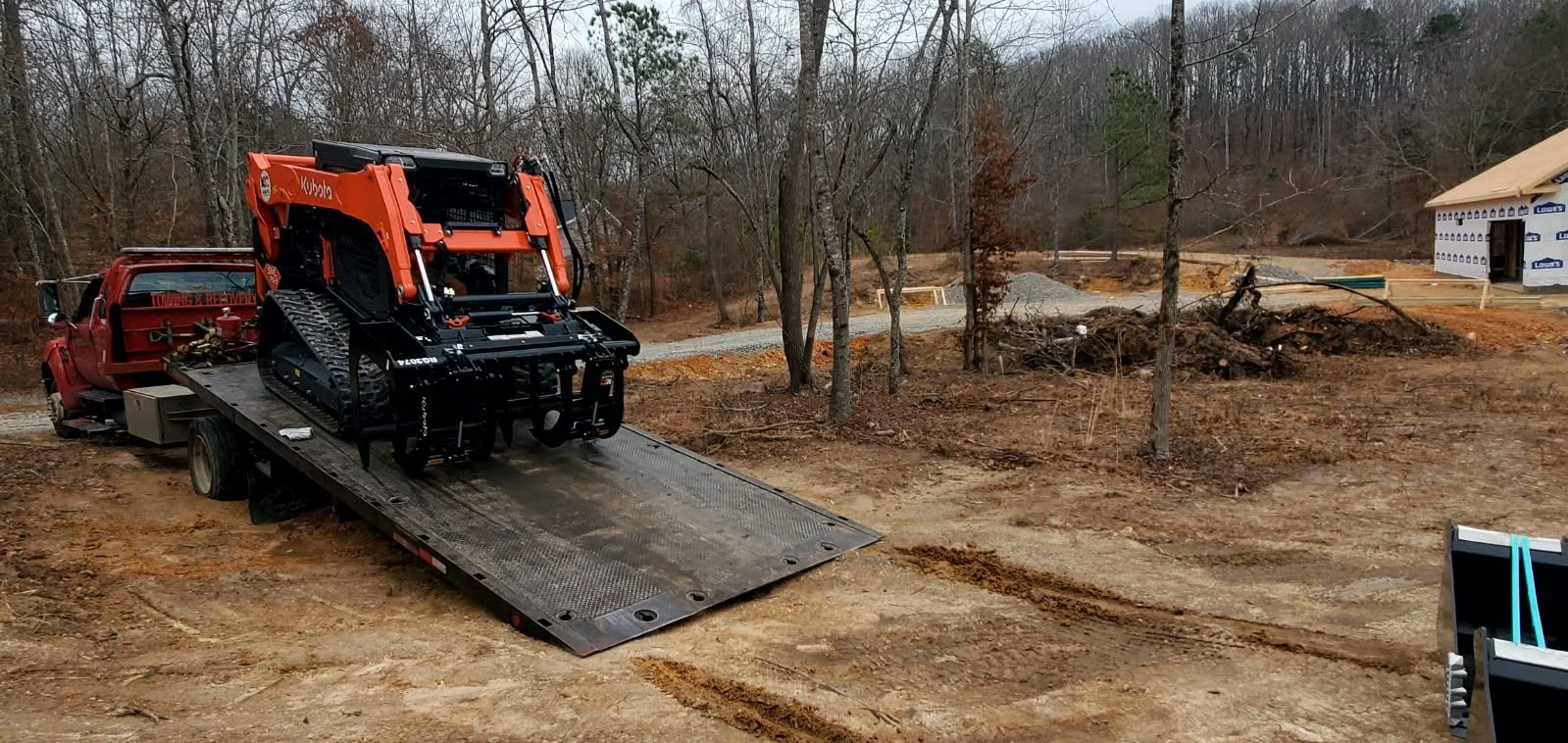 Construction site with a red truck carrying a compact excavator on a flatbed in a wooded area, with a partially built house in the background.