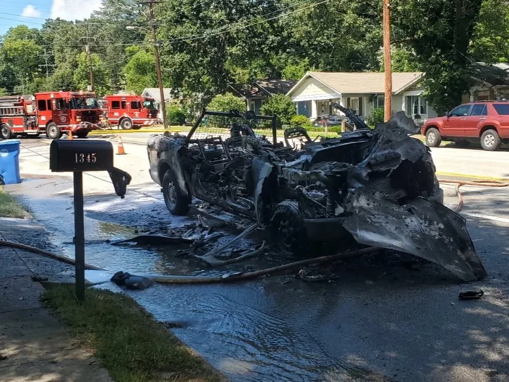 Burnt remains of a vehicle after a fire, with water and hoses around it, near a house and several parked cars, with fire trucks in the background and a tow truck on the way.