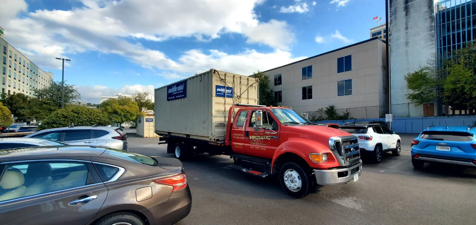 Red moving truck with a large white shipping container on the back, parked among cars in an urban lot under partly cloudy sky.