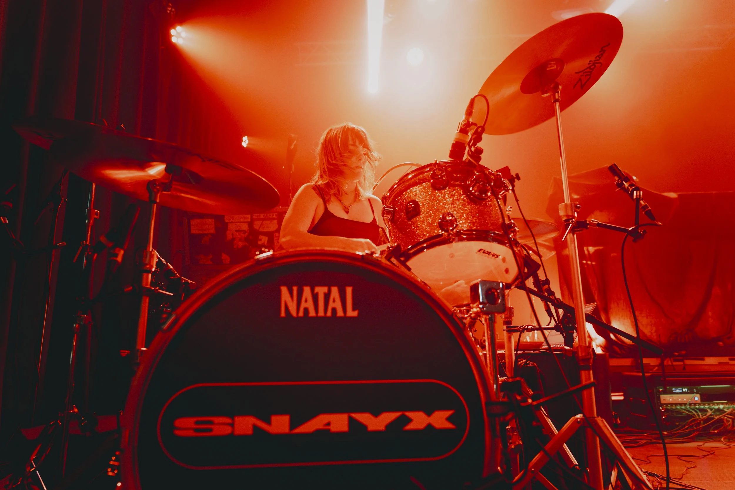 A female drummer playing a drum set with a red and orange stage light illuminating the scene. The bass drum has the word "NATAL" and "SNAXX" on it.