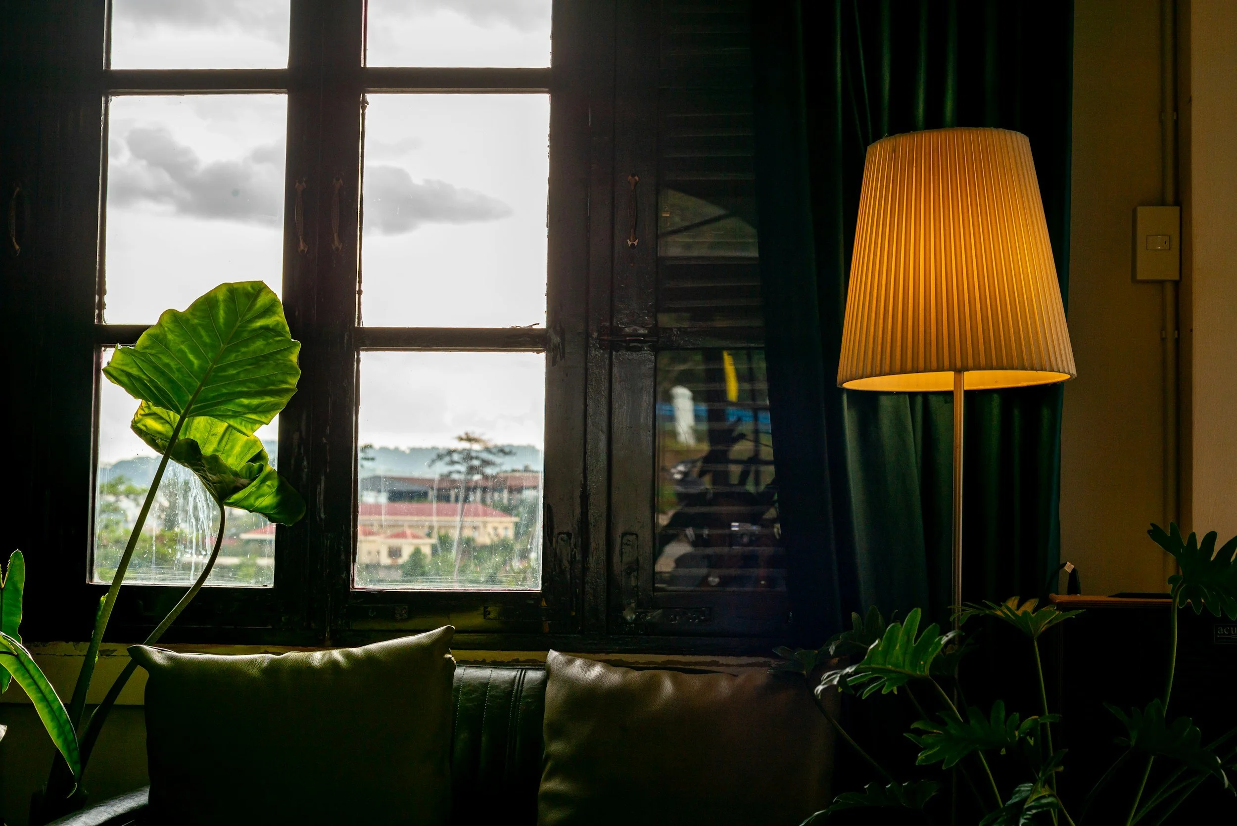 Indoor scene with a window showing a cloudy sky, a tall floor lamp with a pleated shade, green curtains, a large green leaf plant, and part of a dark sofa.