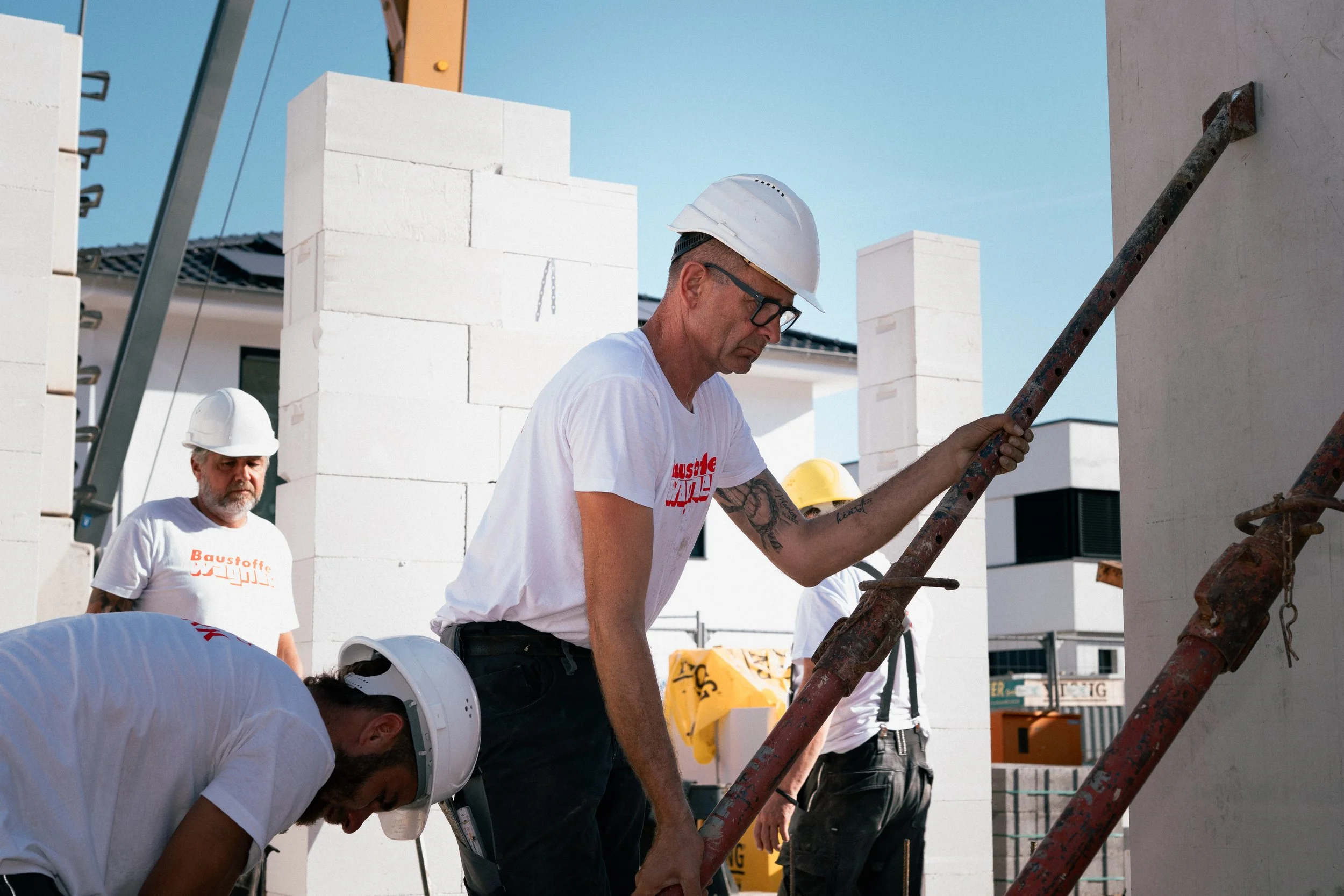 Bauarbeiter bei der Arbeit auf einer Baustelle, sie tragen Helme und helle T-Shirts, einige arbeiten an einer Wand aus weißen Ziegeln, während andere auf einer Gerüststufe stehen.
