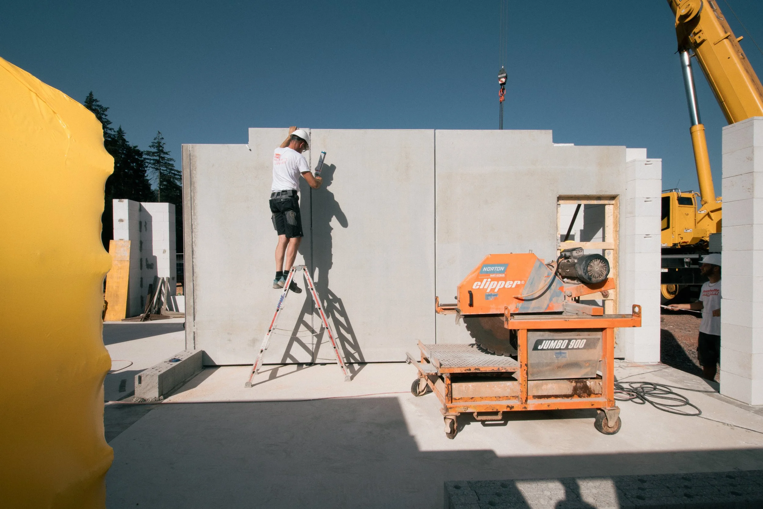 Ein Bauarbeiter steht auf einer Leiter vor einer weißen Wand und liest eine Bauanleitung. Es gibt Baustellengeräte und eine Baustellenumgebung, inklusive Kran und Baucontainer.