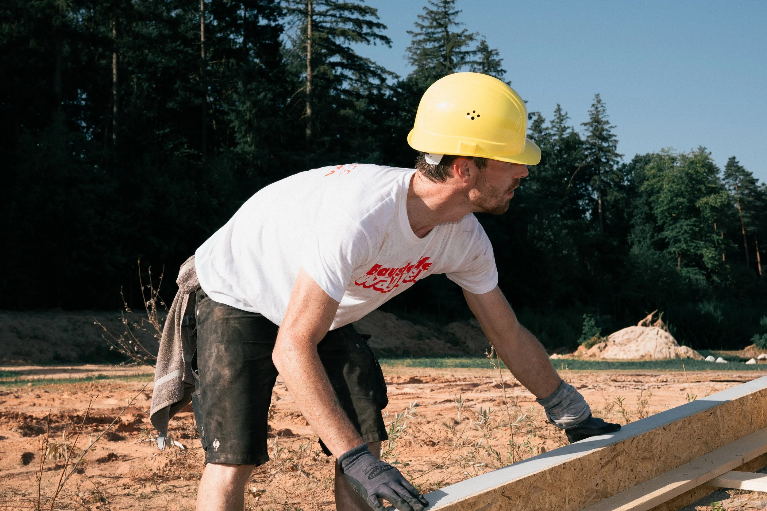 Ein Mann trägt einen gelben Bauhelm und arbeitet auf einer Baustelle im Freien mit Bäumen im Hintergrund.
