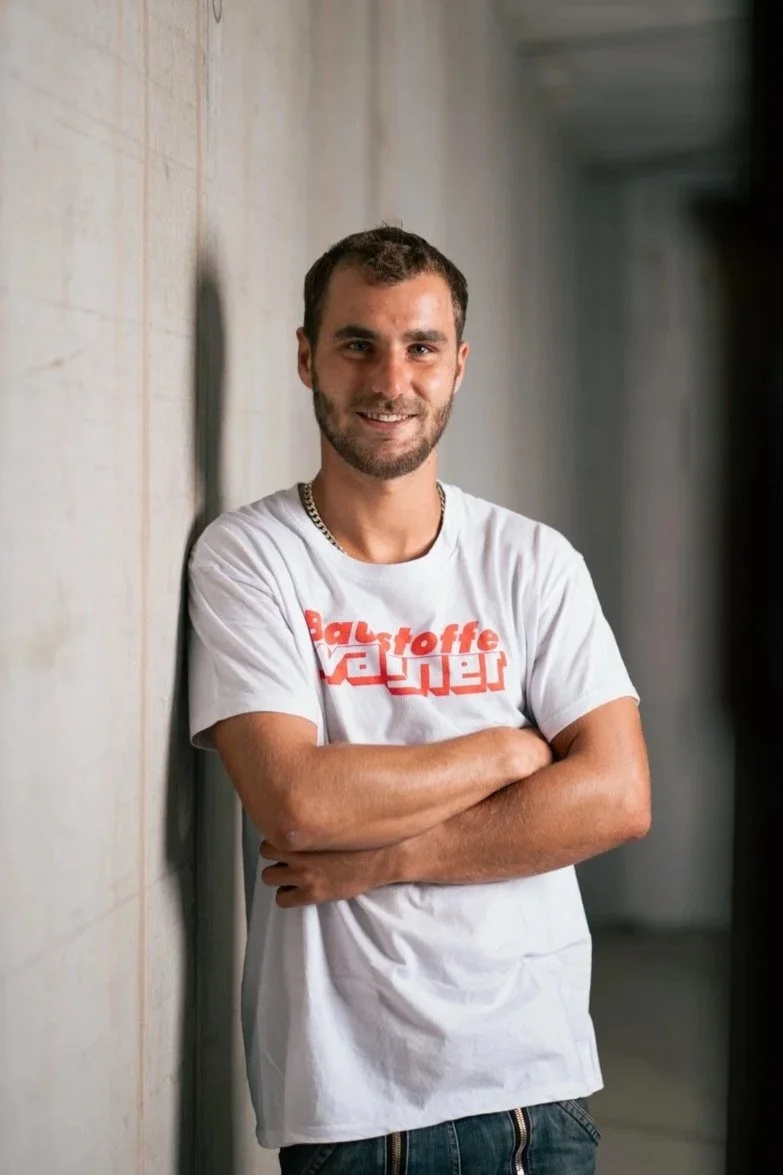 Ein junger Mann mit dunklem Haar und Bart, der an einer Betonwand lehnt und lächelnd in die Kamera schaut, trägt ein weißes T-Shirt mit rotem Logo.