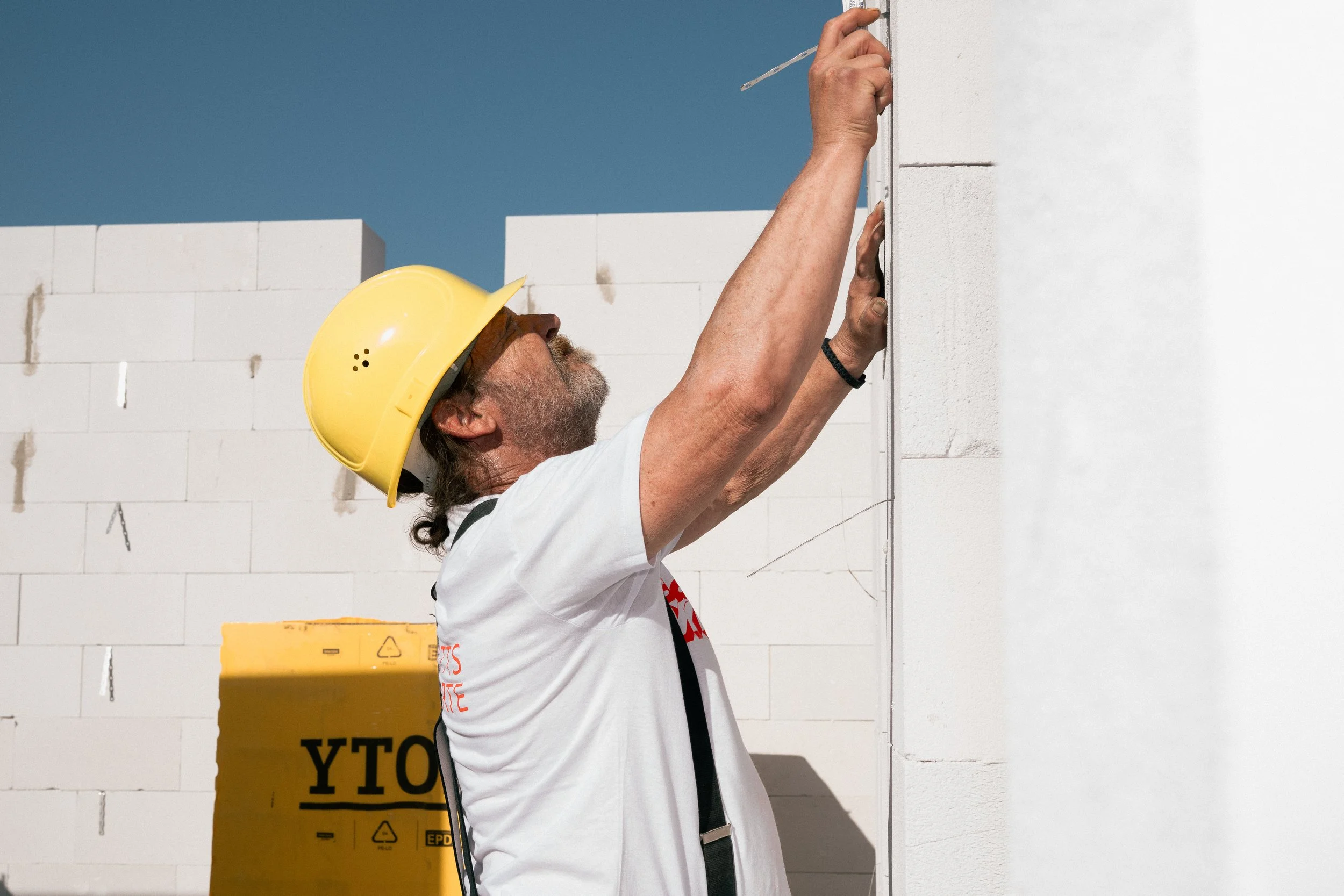 Ein Handwerker trägt einen gelben Schutzhelm und arbeitet an einer weißen Mauer mit einer Wasserwaage.