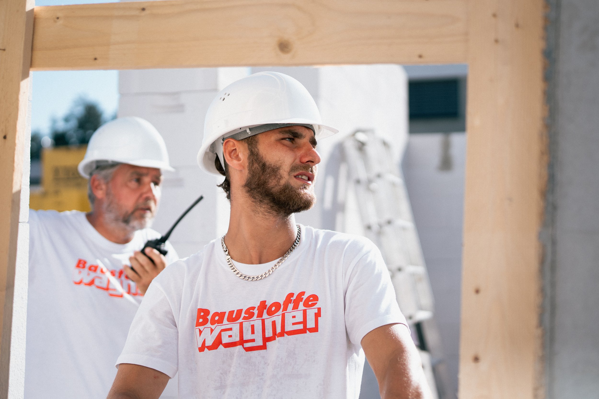 Zwei Männer auf einer Baustelle, einer trägt einen weißen Helm und eine goldene Kette, während er nach oben schaut, der andere trägt ebenfalls einen weißen Helm und hält ein Funkgerät in der Hand.