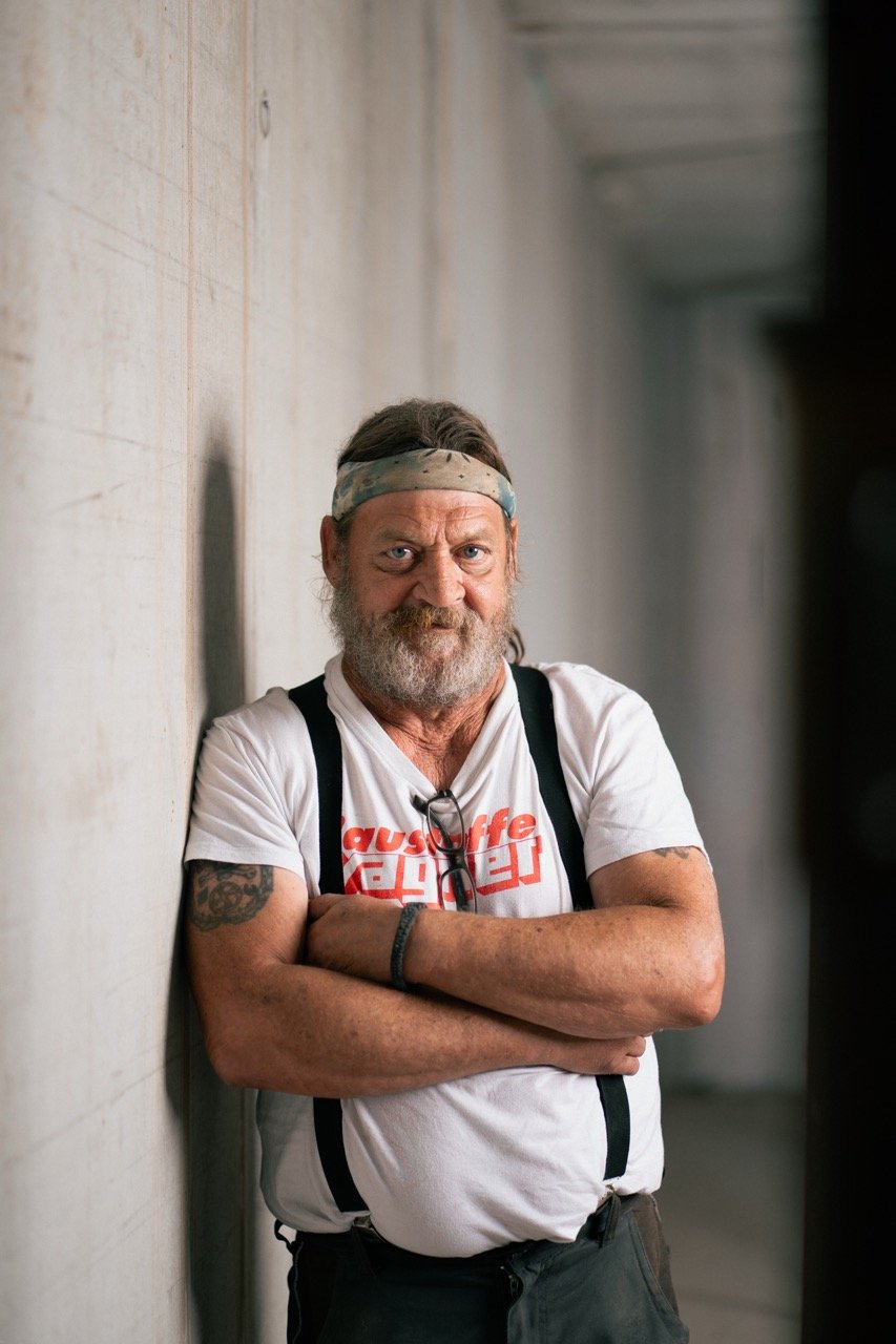 Ein Mann mit grauem Bart, tätowierten Armen und einem bunten Kopfband lehnt an einer Wand. Er trägt ein weißes T-Shirt mit rotem Text und hat eine Brille an der Brust. Er schaut ernst in die Kamera.