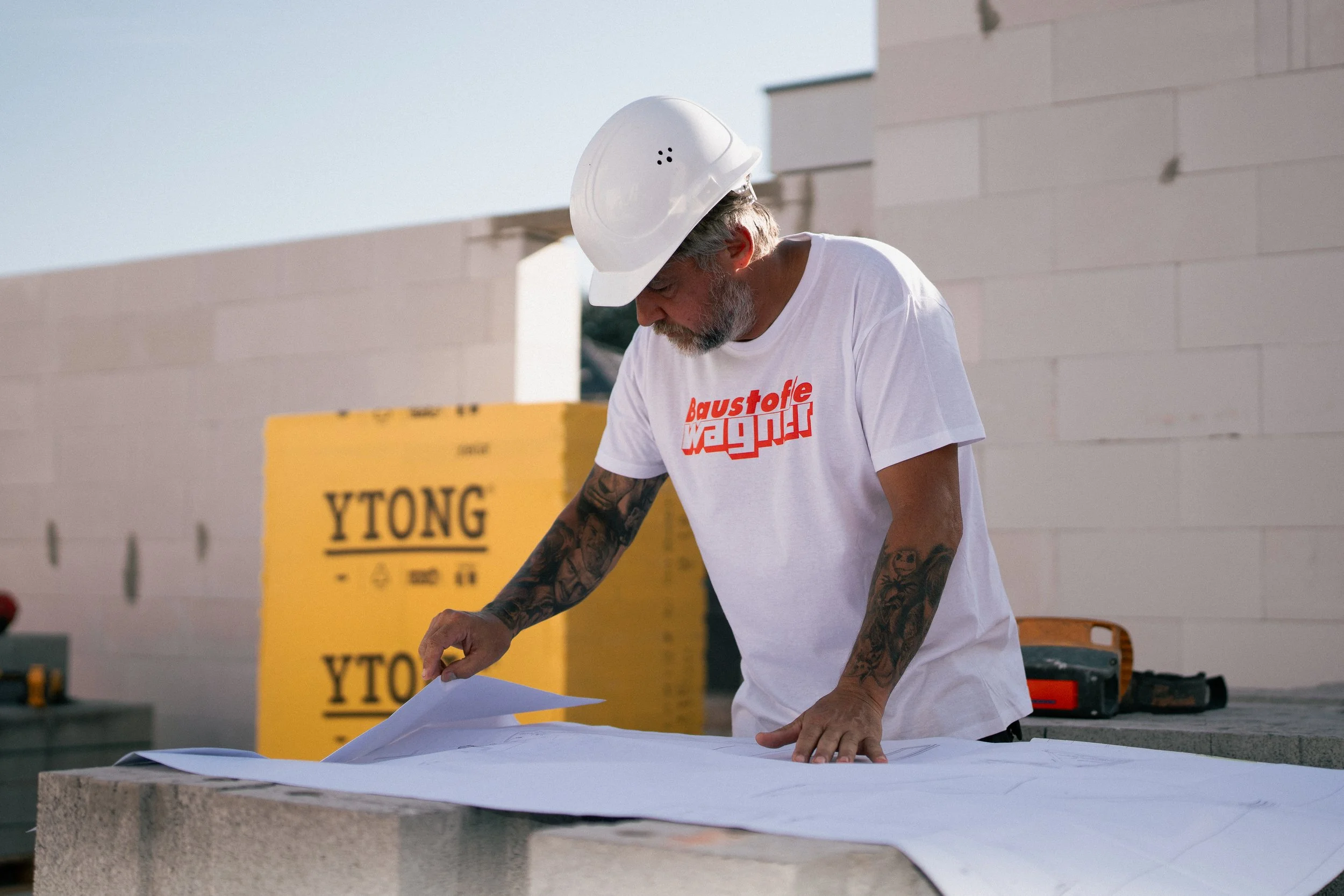 Ein Mann mit grauem Bart, Tattoos an den Armen, trägt ein weißes T-Shirt mit rotem Schriftzug und eine weiße Schutzkappe, arbeitet an einem Bauprojekt im Freien.