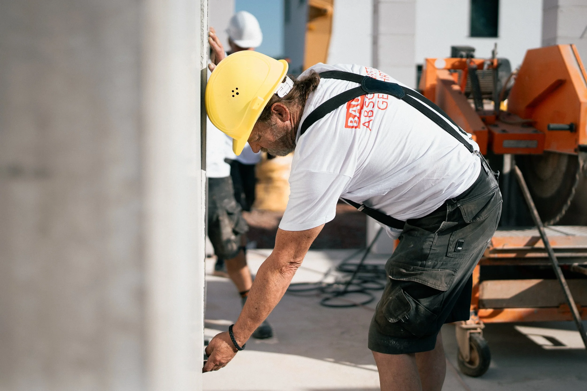 Ein Bauarbeiter trägt einen Schutzhelm und arbeitet an einer Baustelle, er beugt sich vor und inspiziert eine Wand. Im Hintergrund sind Baumaschinen und weitere Arbeiter zu sehen.