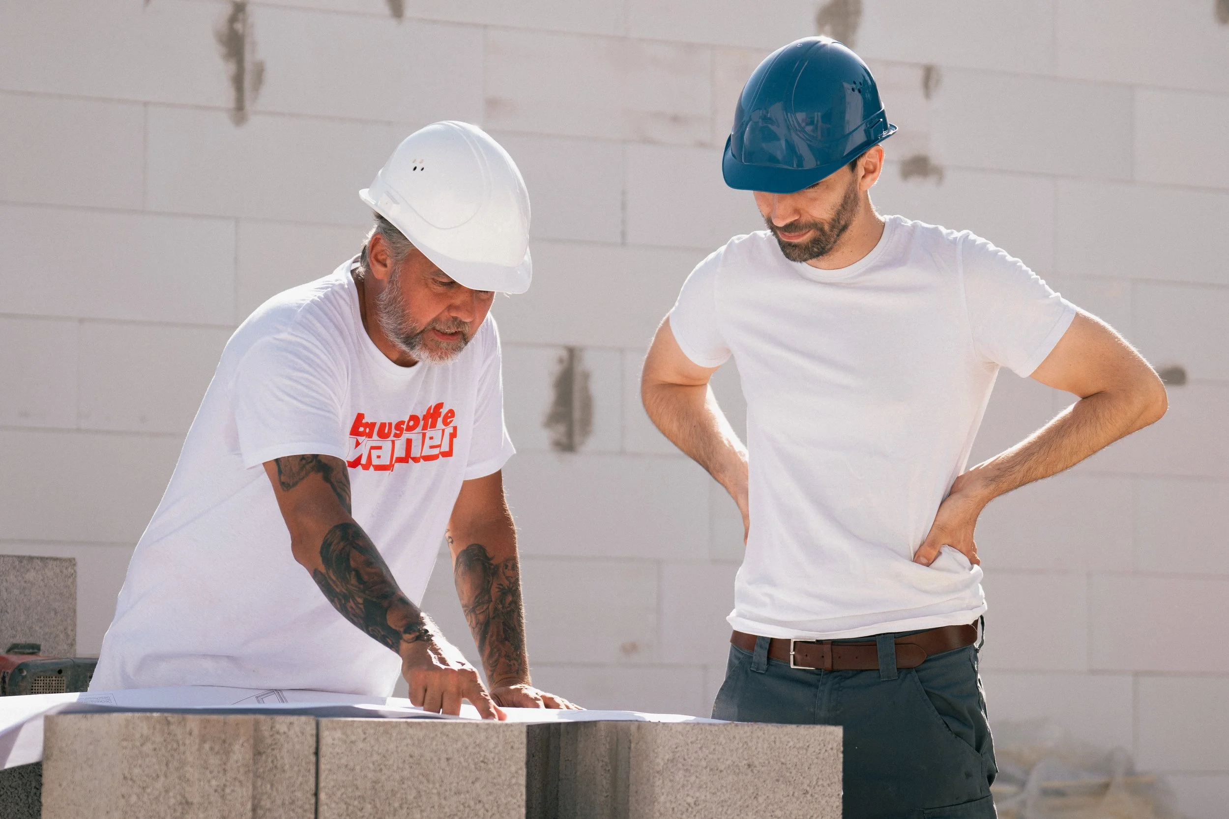 Zwei Männer in Bauhelmen diskutieren auf einer Baustelle über eine Bauzeichnung oder einen Plan.