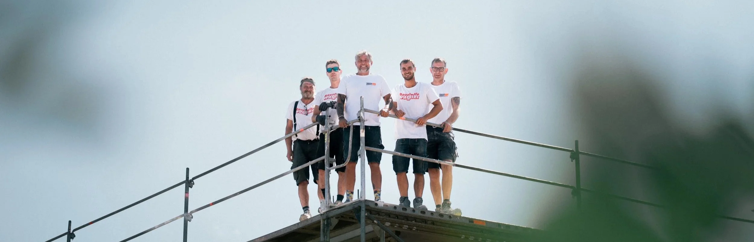 Fünf Männer auf einer Baustellengerüst, alle tragen weiße T-Shirts, einige haben Schweißbänder und Schutzbrillen, lachen und posieren für die Kamera im Freien.