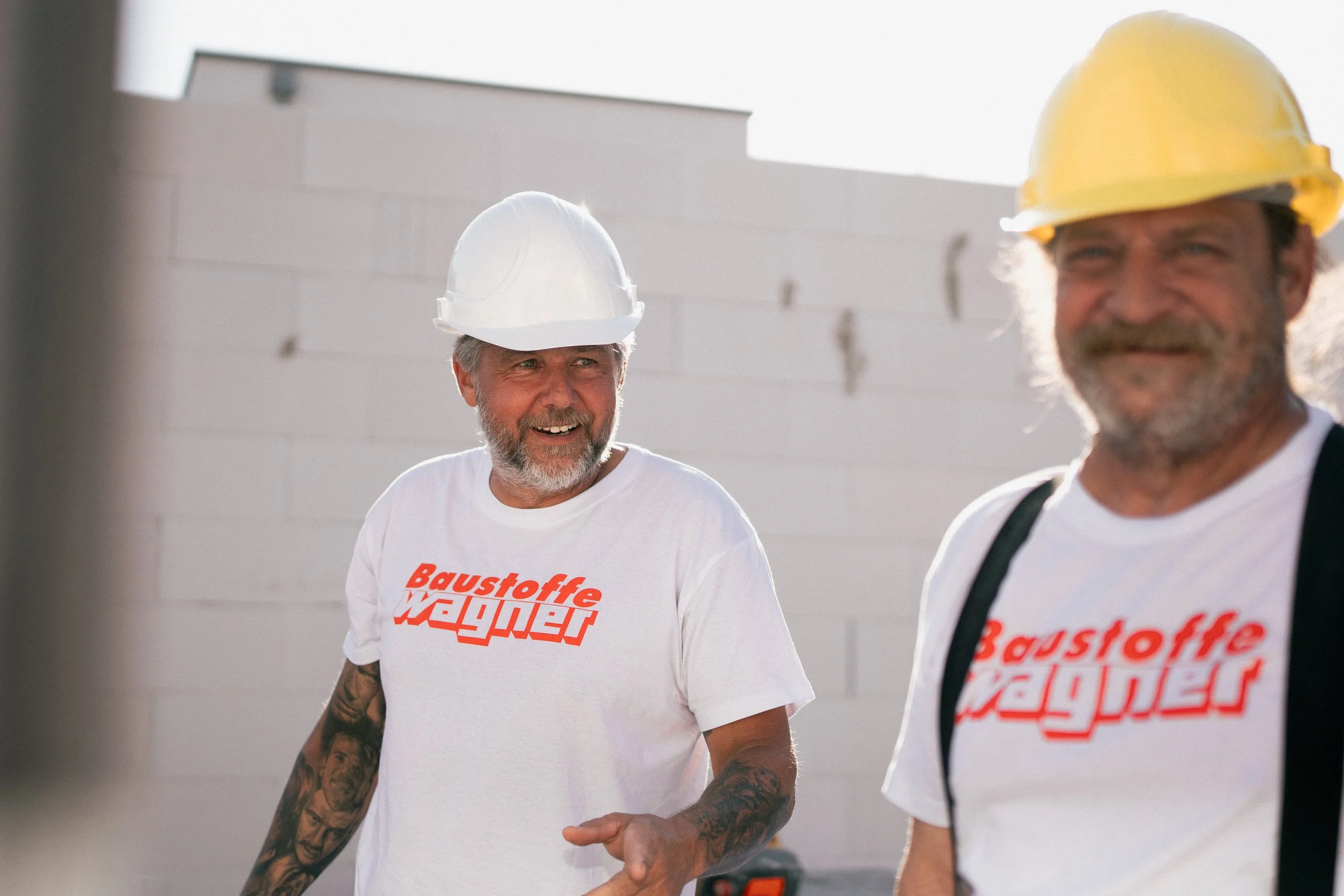 Zwei Männer mit Bauhelmen und T-Shirts mit rotem Aufdruck, die sich auf einer Baustelle unterhalten, im Hintergrund eine weiße Wand.