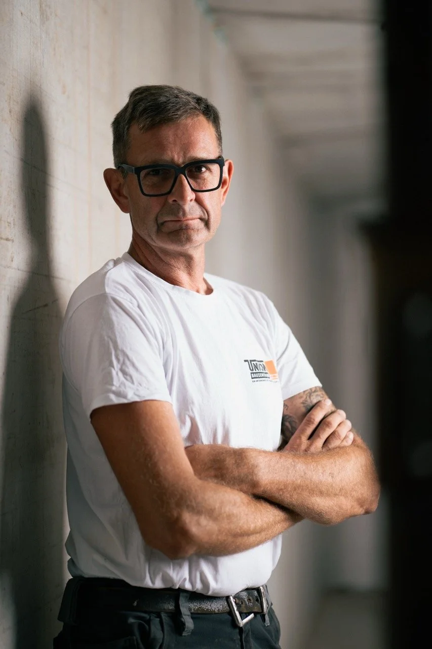 Ein Mann mit Brille lehnt an einer Betonwand, trägt ein weißes T-Shirt und hat den Arm verschränkt.
