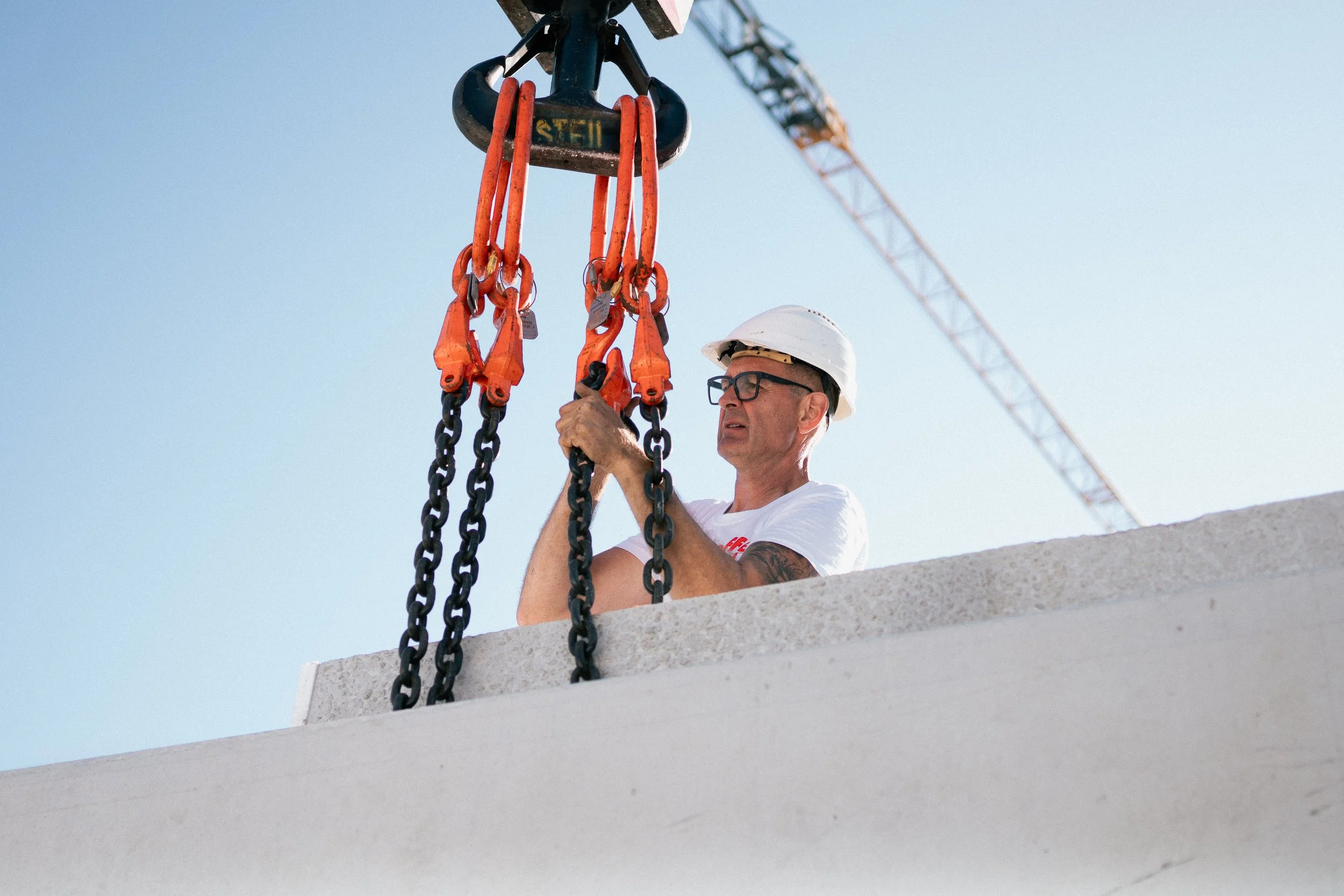 Ein Handwerker trägt Schutzhelm und arbeitet an einem Bauprojekt, während er ein Seilzuggerät hält. Im Hintergrund ist ein Baukran gegen den Himmel sichtbar.
