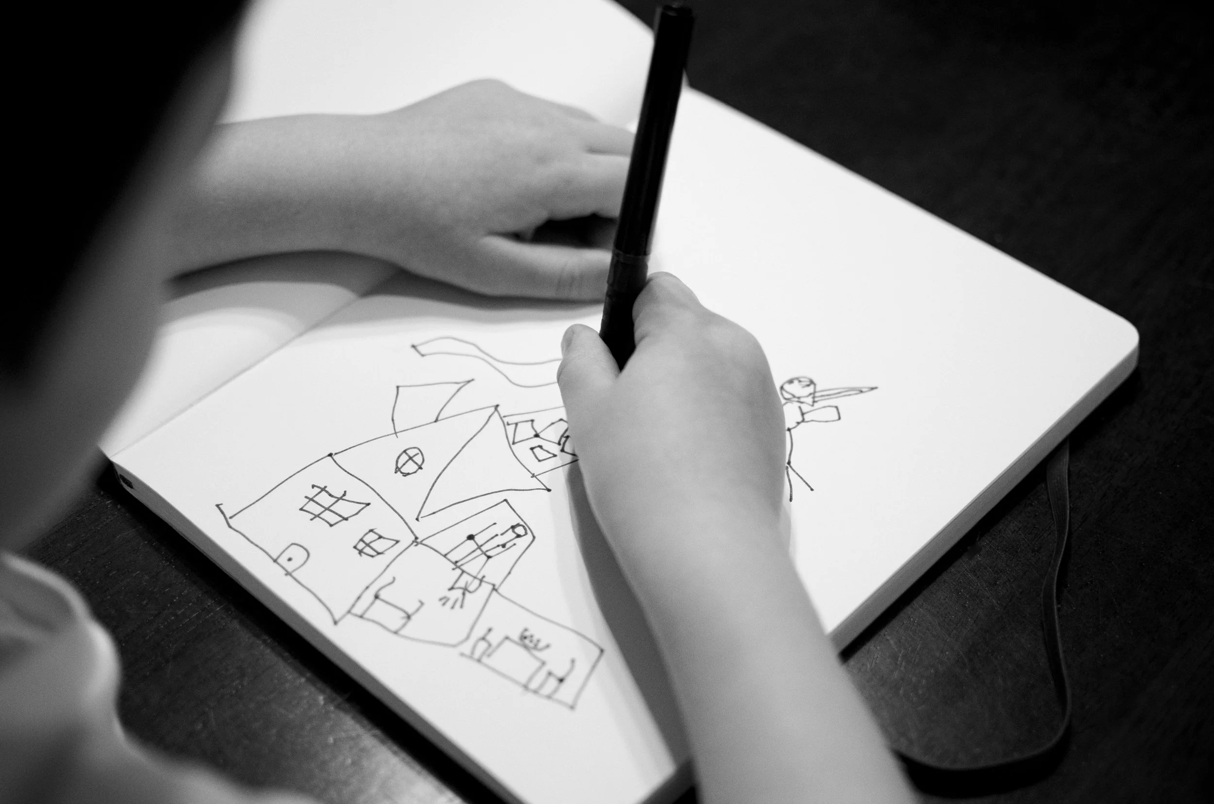 Black and white photo of a child drawing a house and outdoor scene in a sketchbook with a black pen, viewed through a partially blocked camera lens.
