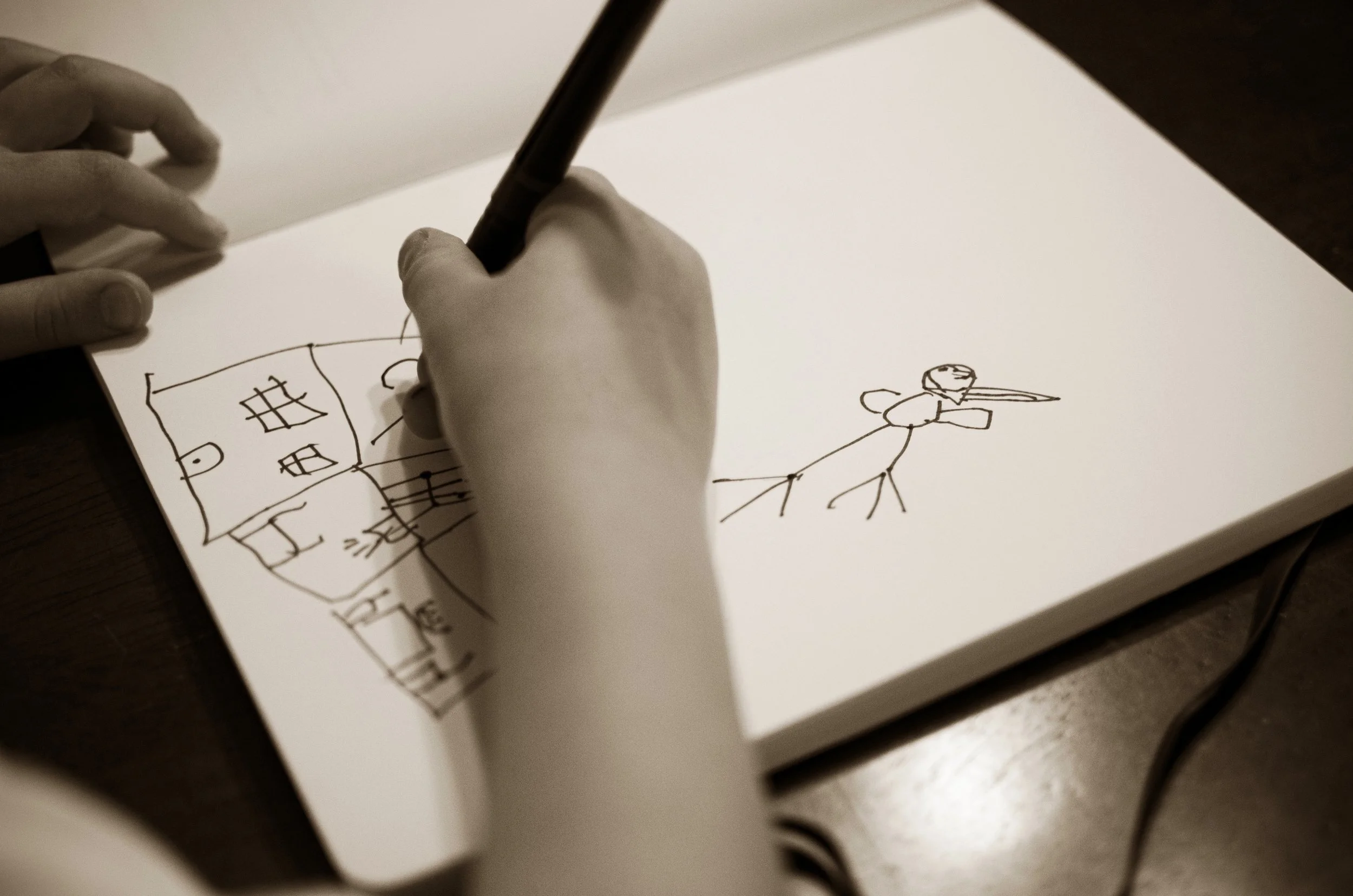 Hand drawing a house and a stick figure on a piece of white paper.