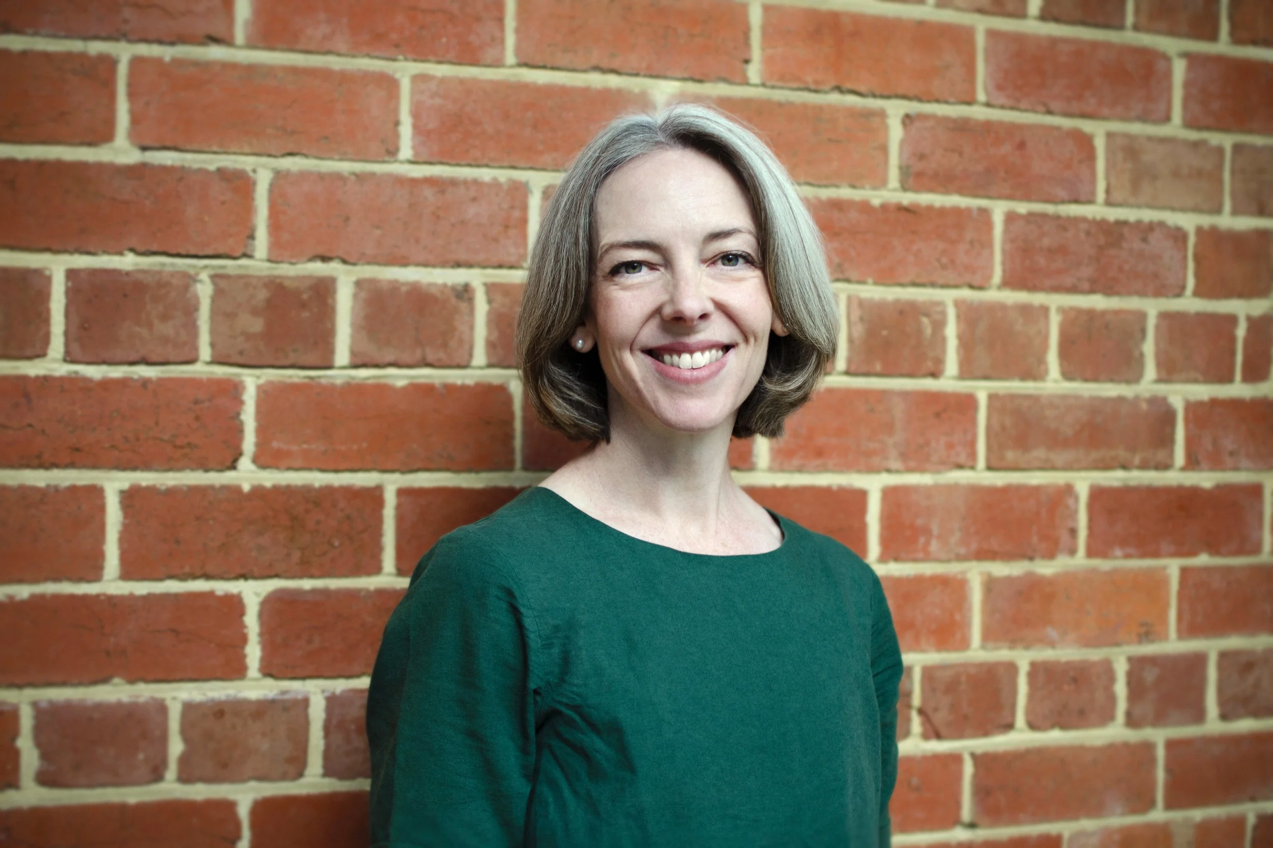 A woman with shoulder-length gray hair smiling next to a red brick wall, wearing a dark green top.