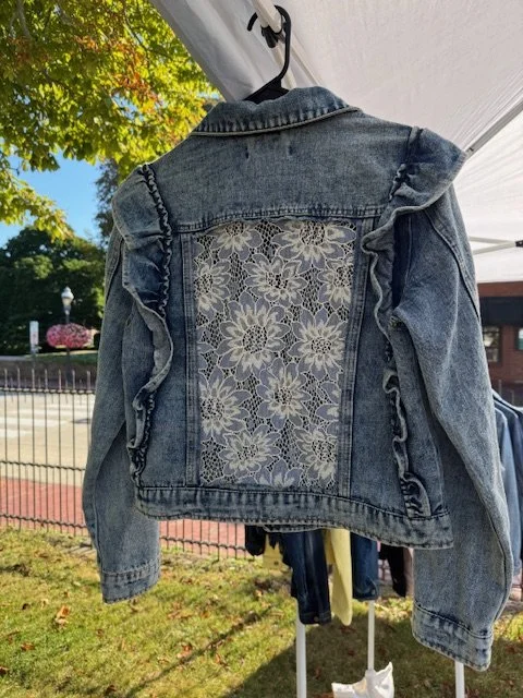 Denim jacket with lace floral back detail and ruffled shoulder accents hanging outdoors.