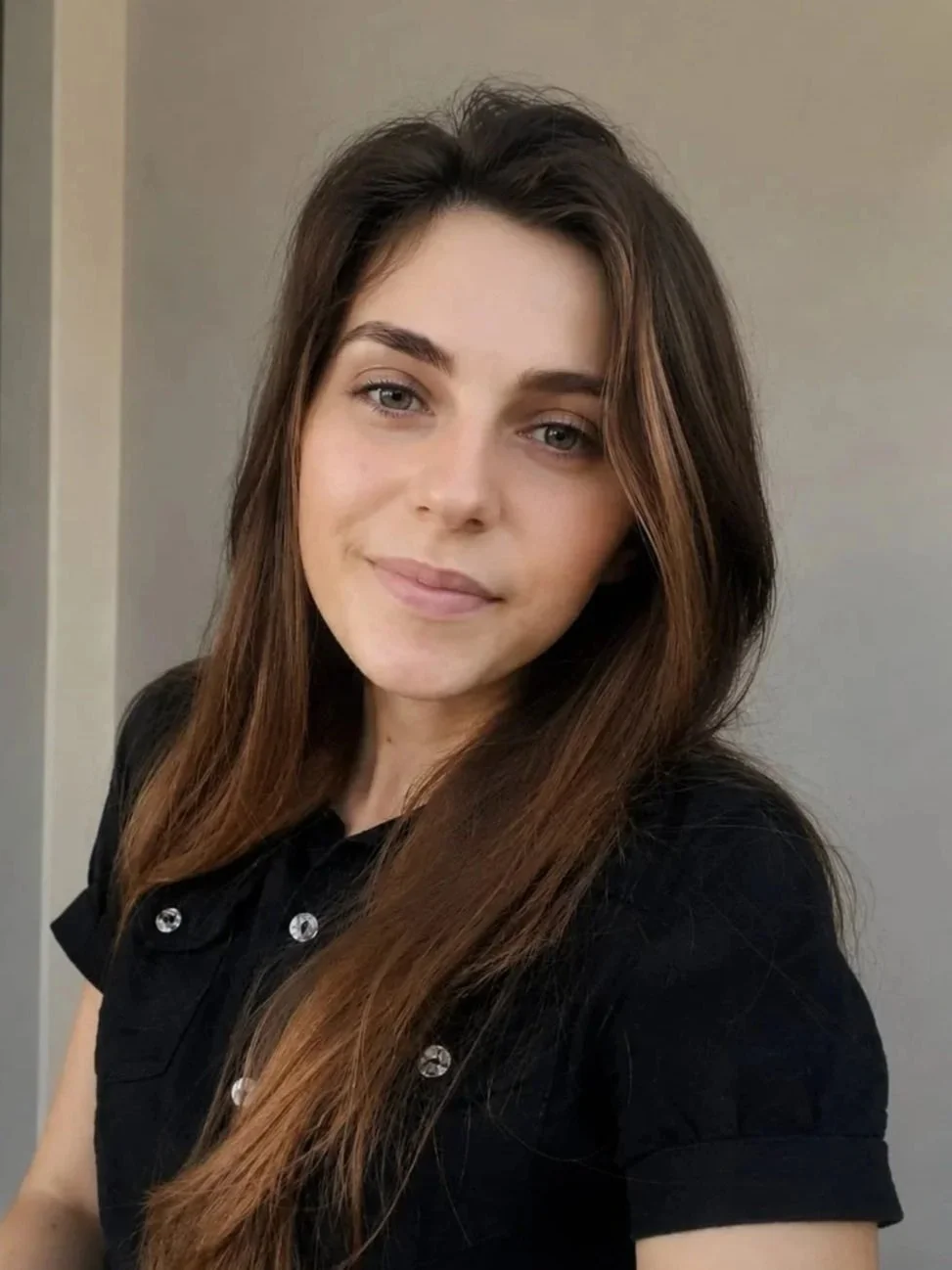 A young woman with long brown hair and blue eyes, wearing a black shirt, is smiling softly at the camera.