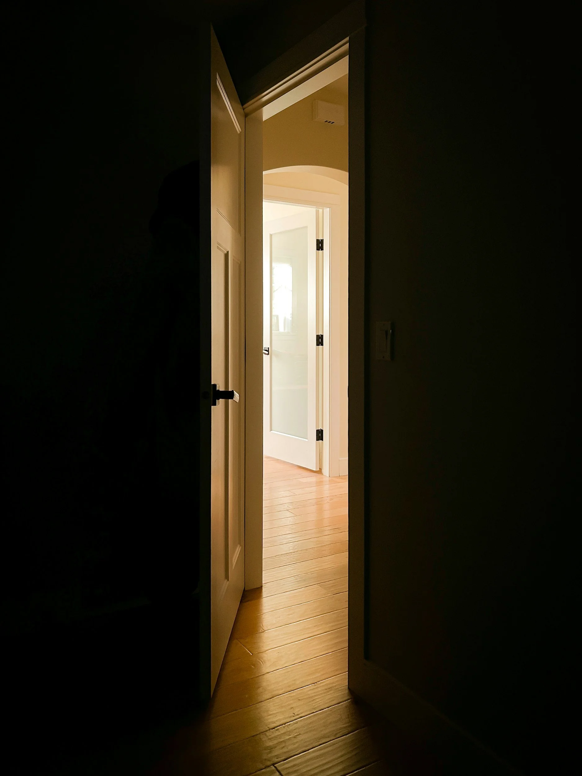 Open door revealing a sunlit room with hardwood flooring and a window in the background.
