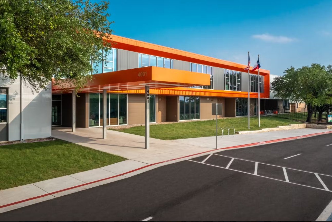Exterior of Norman-Sims Elementary School, a Best Spaces certified project, featuring a two-story building with orange accents, large windows, a landscaped entrance, and a paved drive.