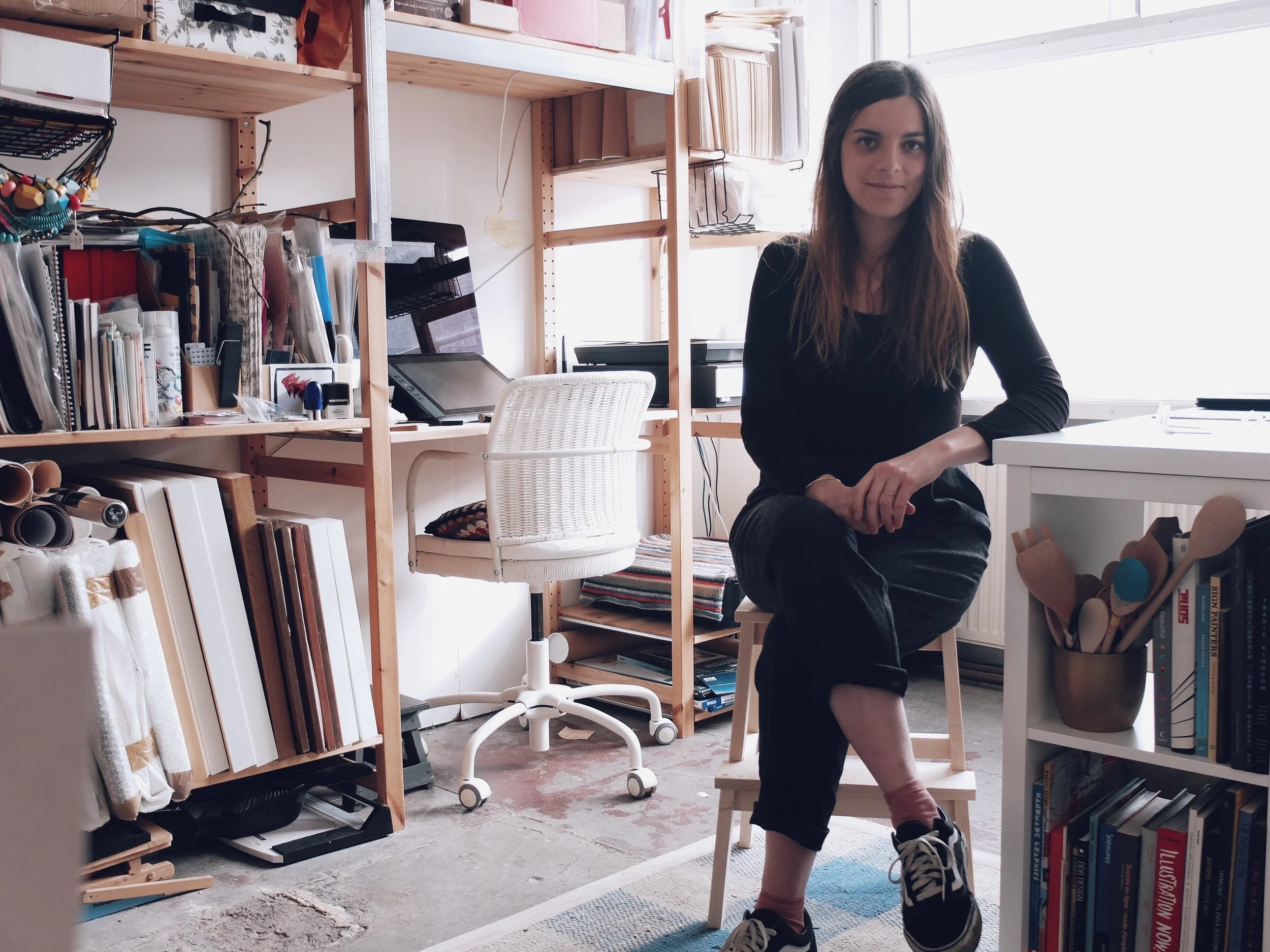 A woman with long brown hair sitting on a small stool in a room filled with books, shelves, and art supplies. She is wearing a black long-sleeve top, black pants, and sneakers, with one leg crossed over the other.