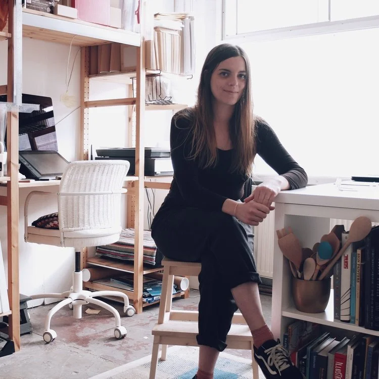 A woman with long brown hair, wearing a black dress, sits on a wooden stool in a room with bookshelves and a white desk, next to a large window.