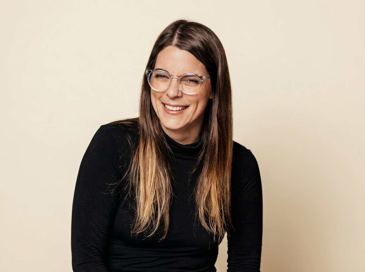 A woman with long brown hair, glasses, and a black turtleneck smiling in front of a plain background.