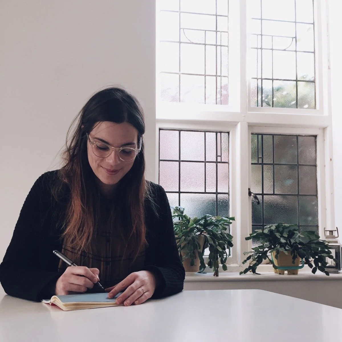 A woman sitting at a white table, writing in a notebook with a pen, near a large window with frosted and clear glass panes, and potted plants on the windowsill.