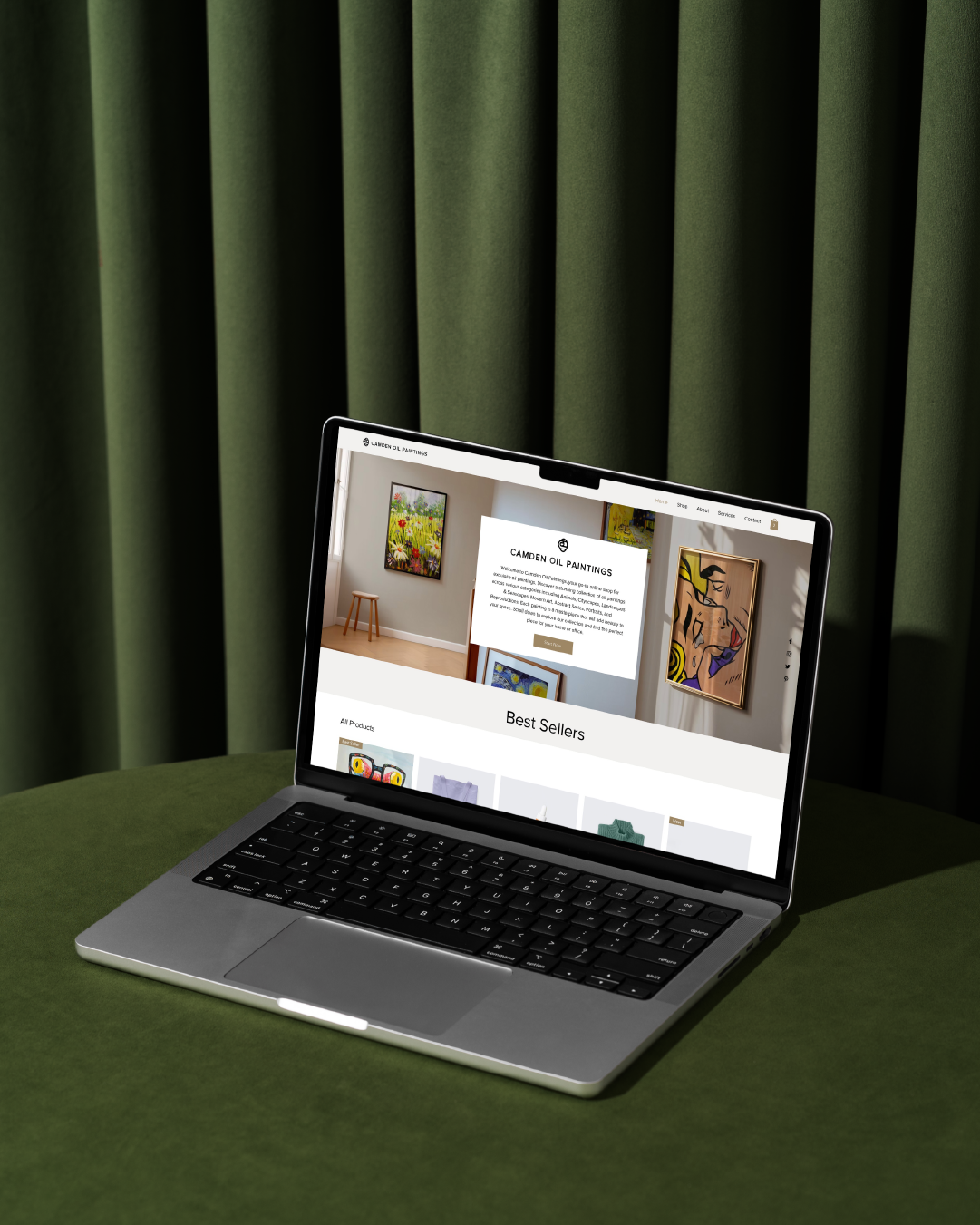 Laptop on a green table displaying the homepage of Camden Oil Paintings website, featuring artwork and navigation menu.
