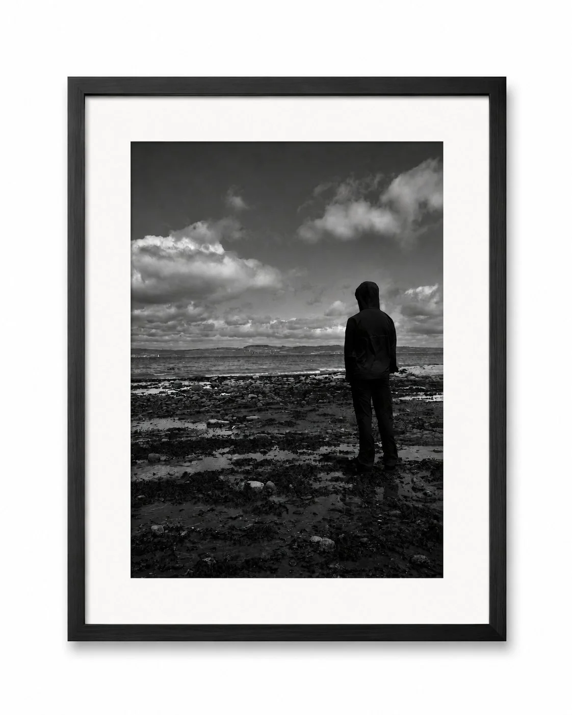 A person standing on a rocky beach looking at the ocean under cloudy skies, in black and white.