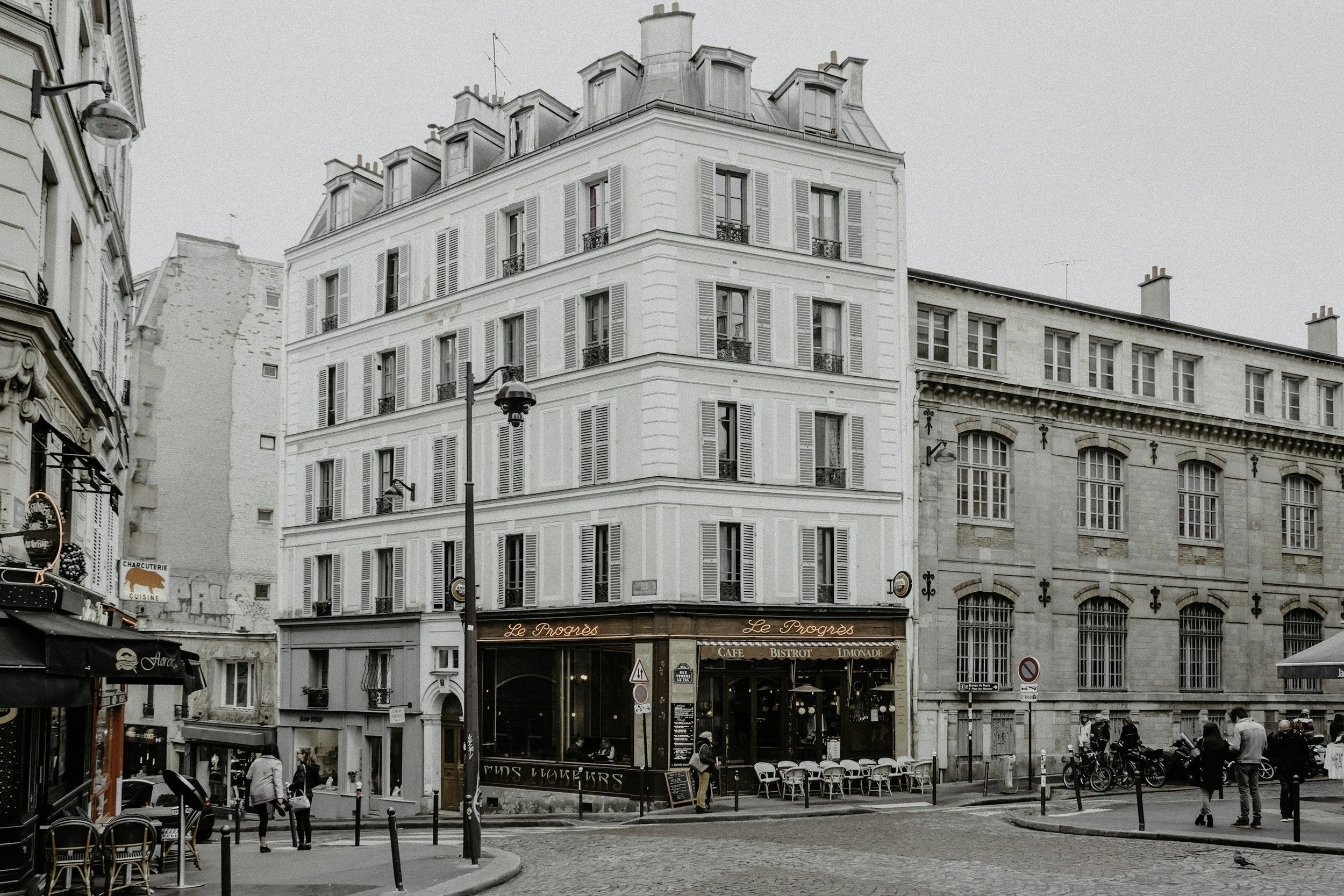 A city street corner with a corner building, people walking, bicycles parked, and a cafe named 'Le Progres' with outdoor seating. French travel