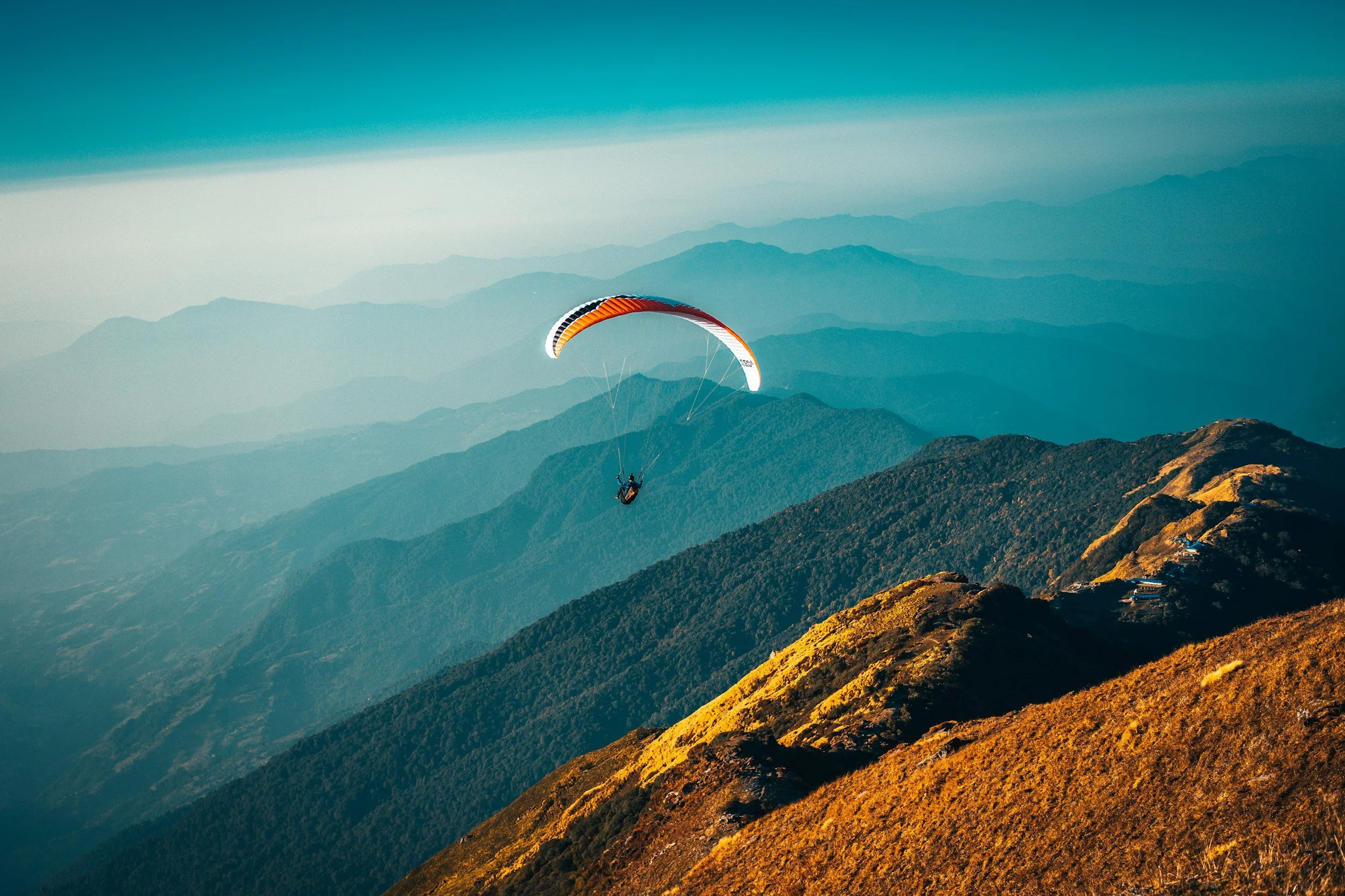 A person paragliding over mountain ridges during sunset, with colorful sky and layered mountainous landscape in the background. Adventure travel