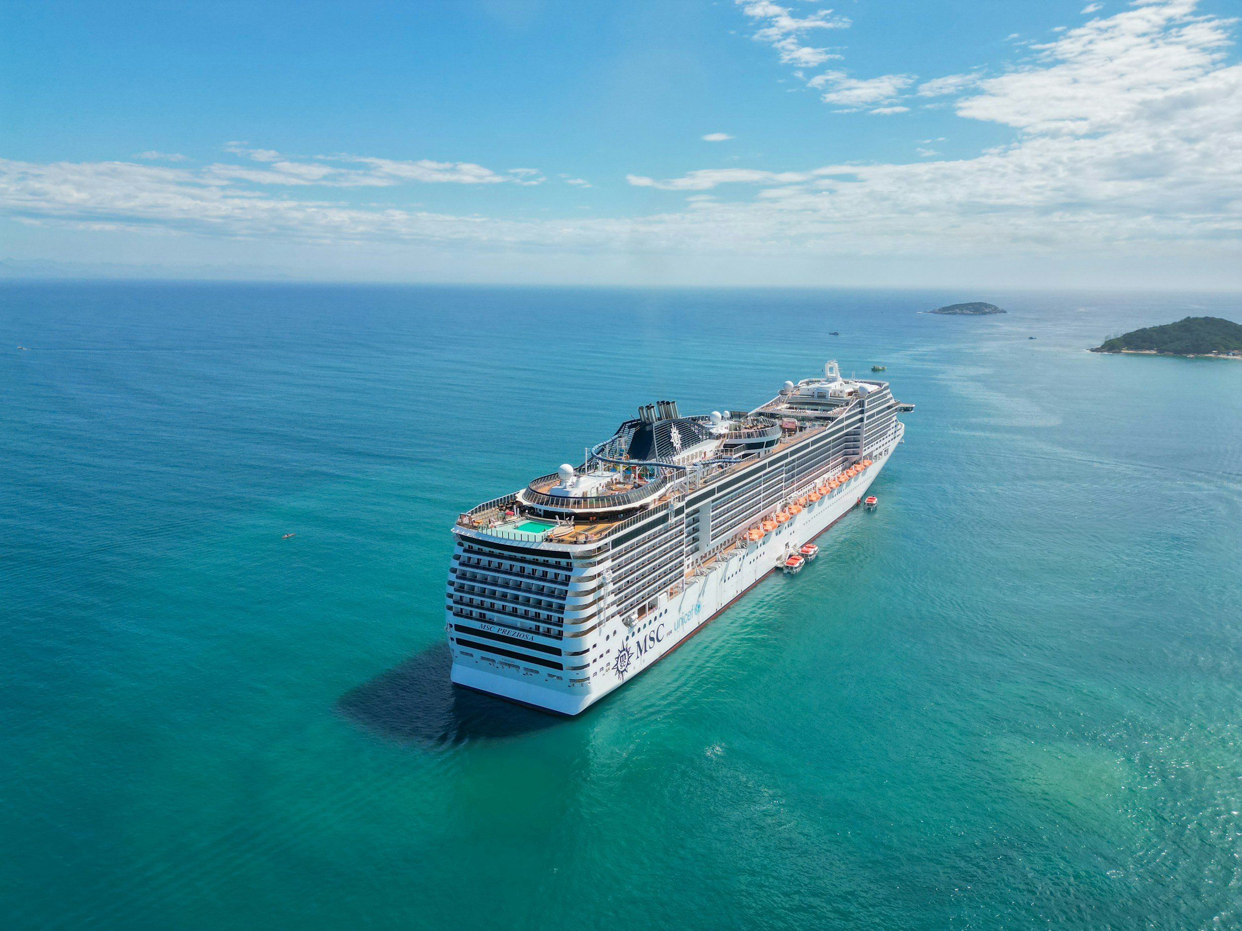 A large white cruise ship with multiple decks sailing in clear blue ocean water near green islands, under a partly cloudy sky.