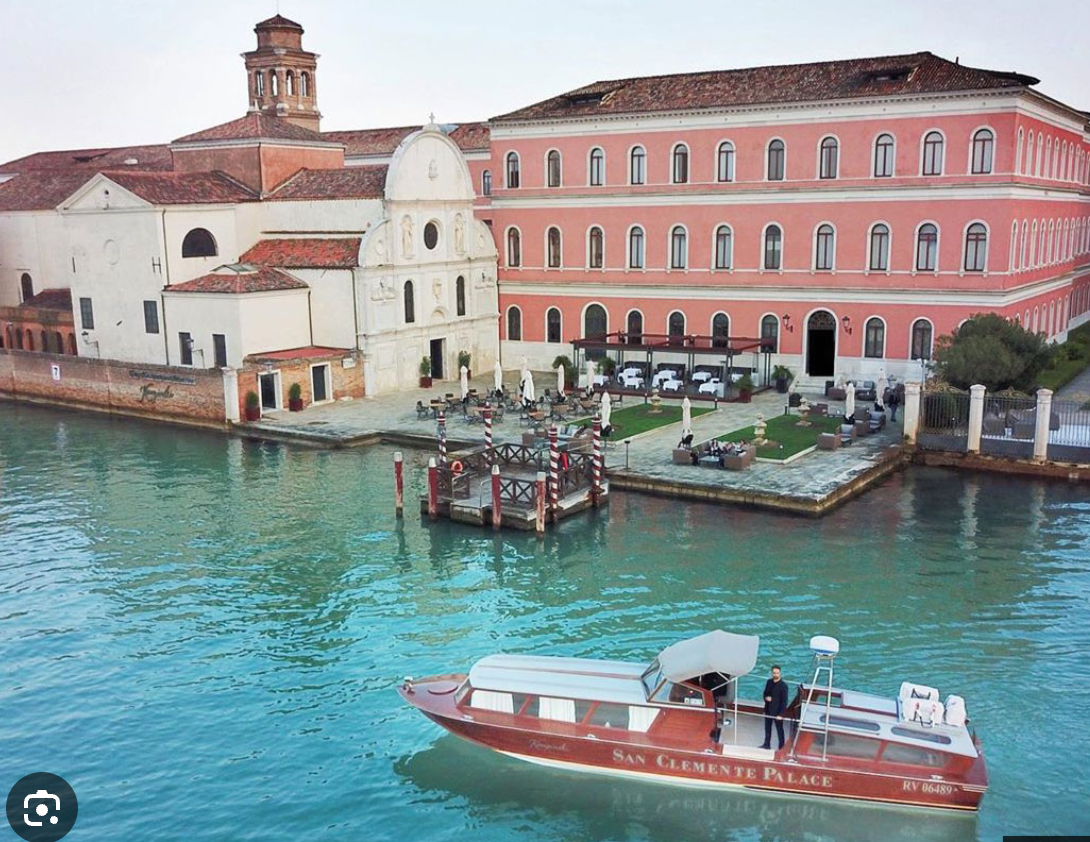 A boat on water near a historic building with a pink facade and white accents, outdoor seating with umbrellas, and a small dock with red and white striped poles.