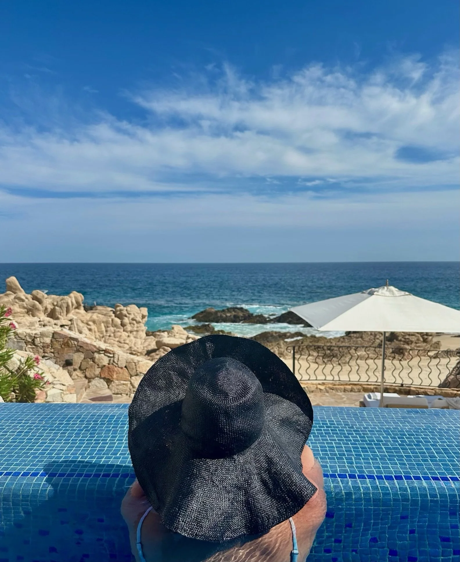 View of a person relaxing in a swimming pool with a large sun hat on their head, overlooking a scenic ocean view with a rocky shoreline, blue sky, and clouds, while sitting on a tiled pool edge under a white umbrella. Caribbean travel