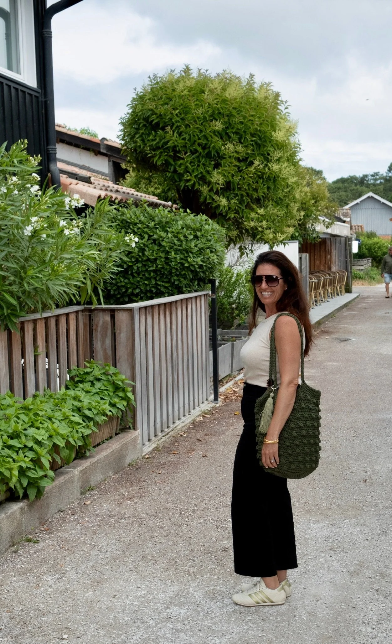 A woman with long brown hair, wearing sunglasses, a light beige sleeveless top, black pants, white sneakers, and carrying a green crochet bag with tassels, standing on a gravel path next to a wooden fence with plants and trees in the background, under a cloudy sky.